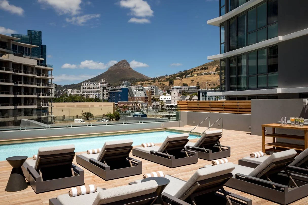 Rooftop swimming pool area with lounge chairs and a cityscape background featuring buildings and a mountain.