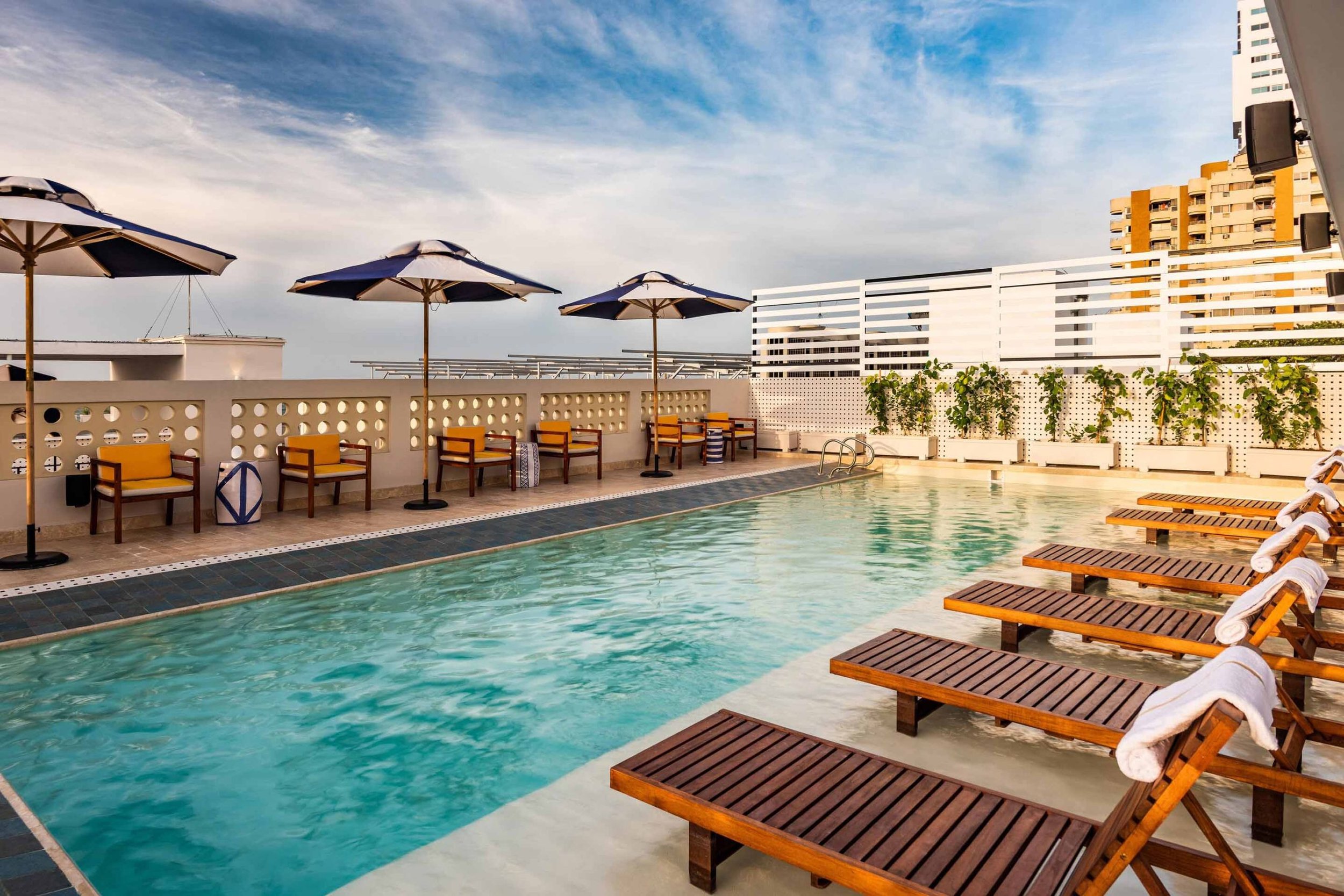 Rooftop swimming pool with wooden lounge chairs and umbrellas, surrounded by a white decorative wall and high-rise buildings in the background under a partly cloudy sky.