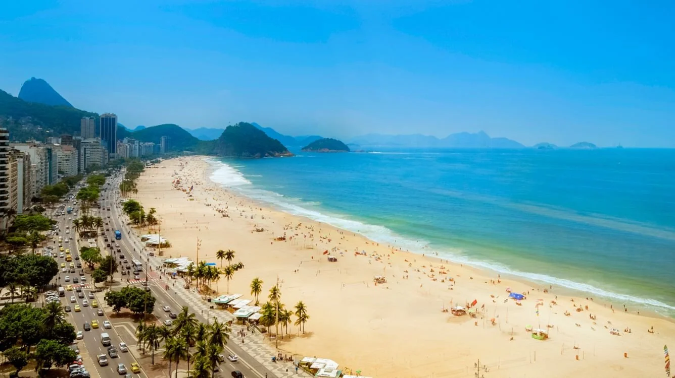 Aerial view of a sunny beach with golden sand, people relaxing, colorful umbrellas, and a blue ocean with gentle waves. High-rise buildings and green mountains are in the background, with traffic on a nearby road.