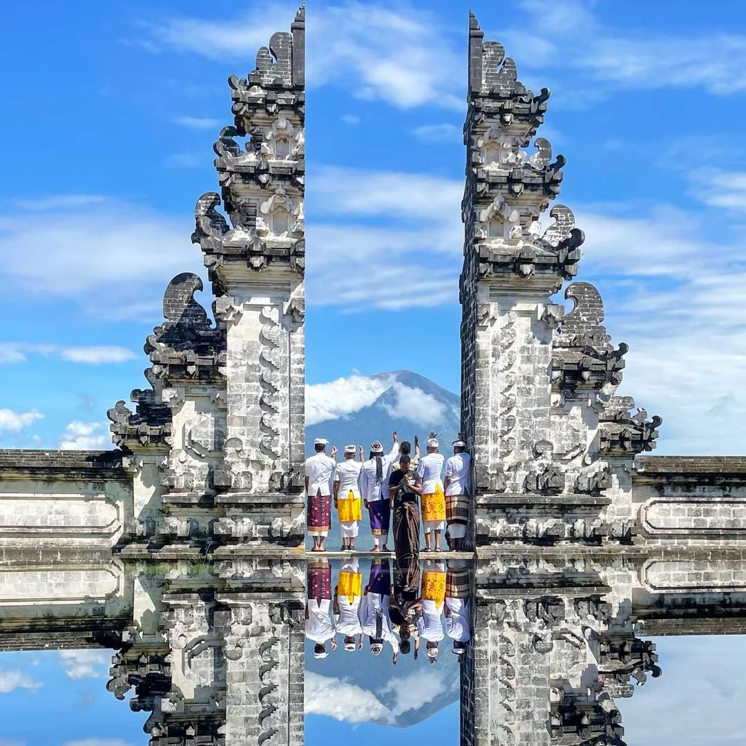 Group of Balinese people in traditional clothing standing between ornate stone gates, with Mount Agung in the background and a clear blue sky.