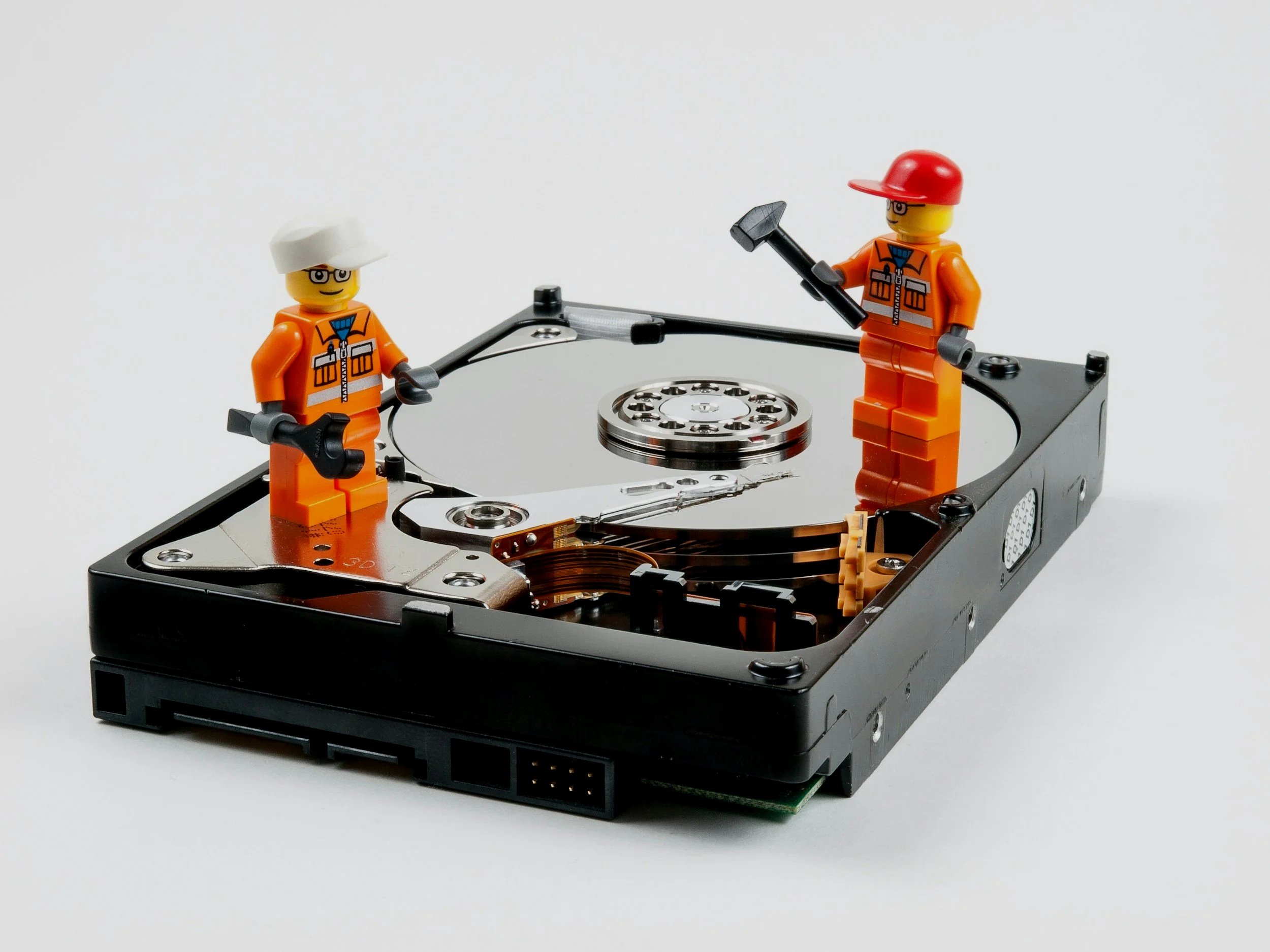 Two LEGO construction worker minifigures standing on an opened computer hard drive with a white background.