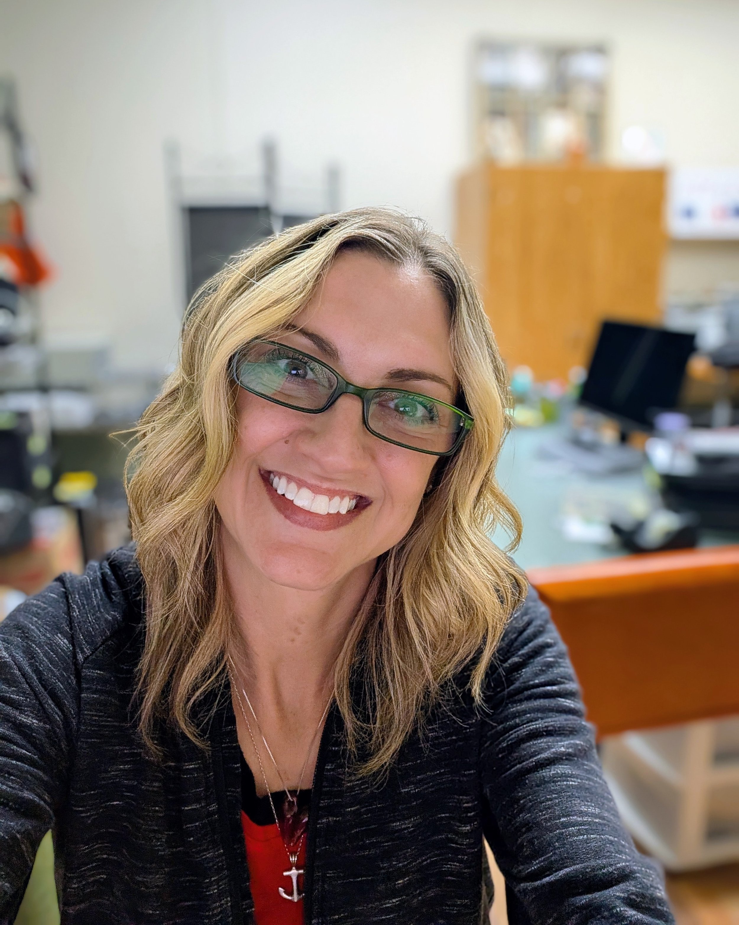 Close-up of a smiling woman with blonde hair and glasses, wearing a black and gray top and a red shirt with a white anchor necklace, in a room with shelves and a computer in the background.