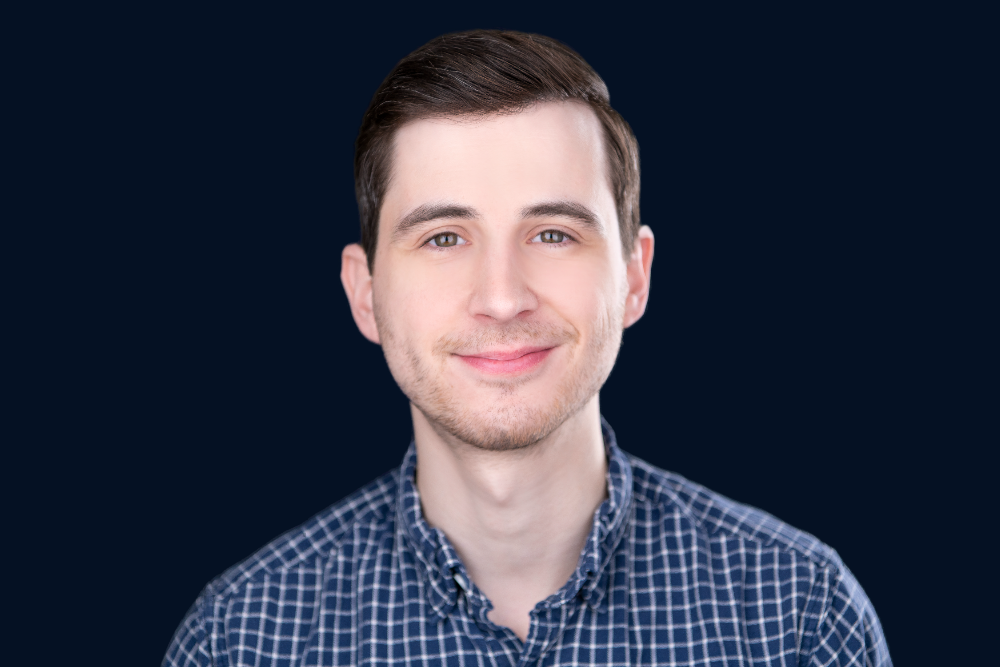 Close-up portrait of a young man with short brown hair and light skin, smiling, wearing a checkered shirt, against a dark background.