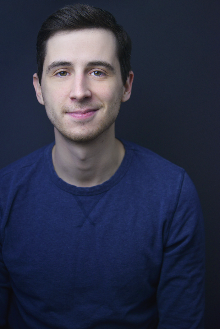A young man with short dark hair, light skin, and a slight smile wearing a navy blue shirt, posing against a dark background.