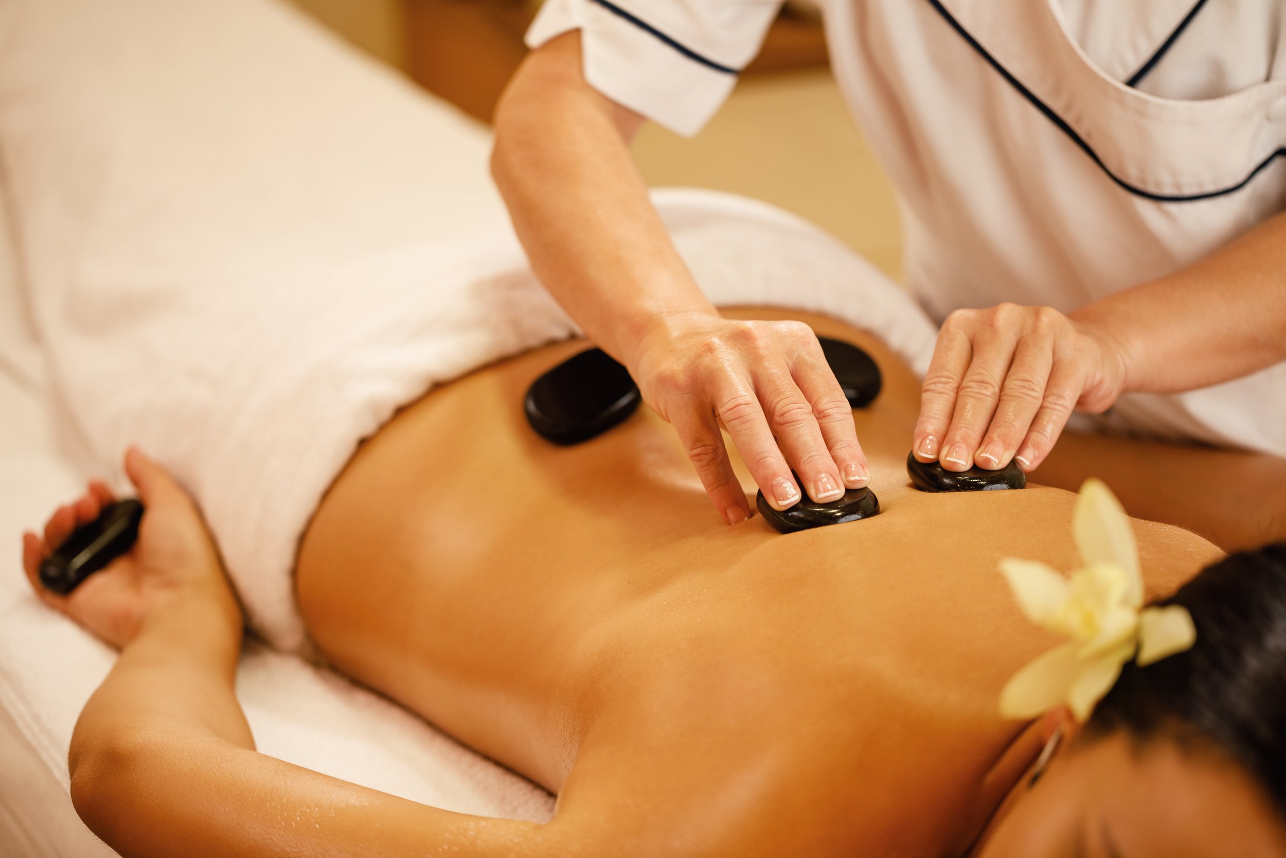 A person is receiving a hot stone massage on their back, with several smooth black stones placed along their spine, while another person is using their hands to massage the stones.