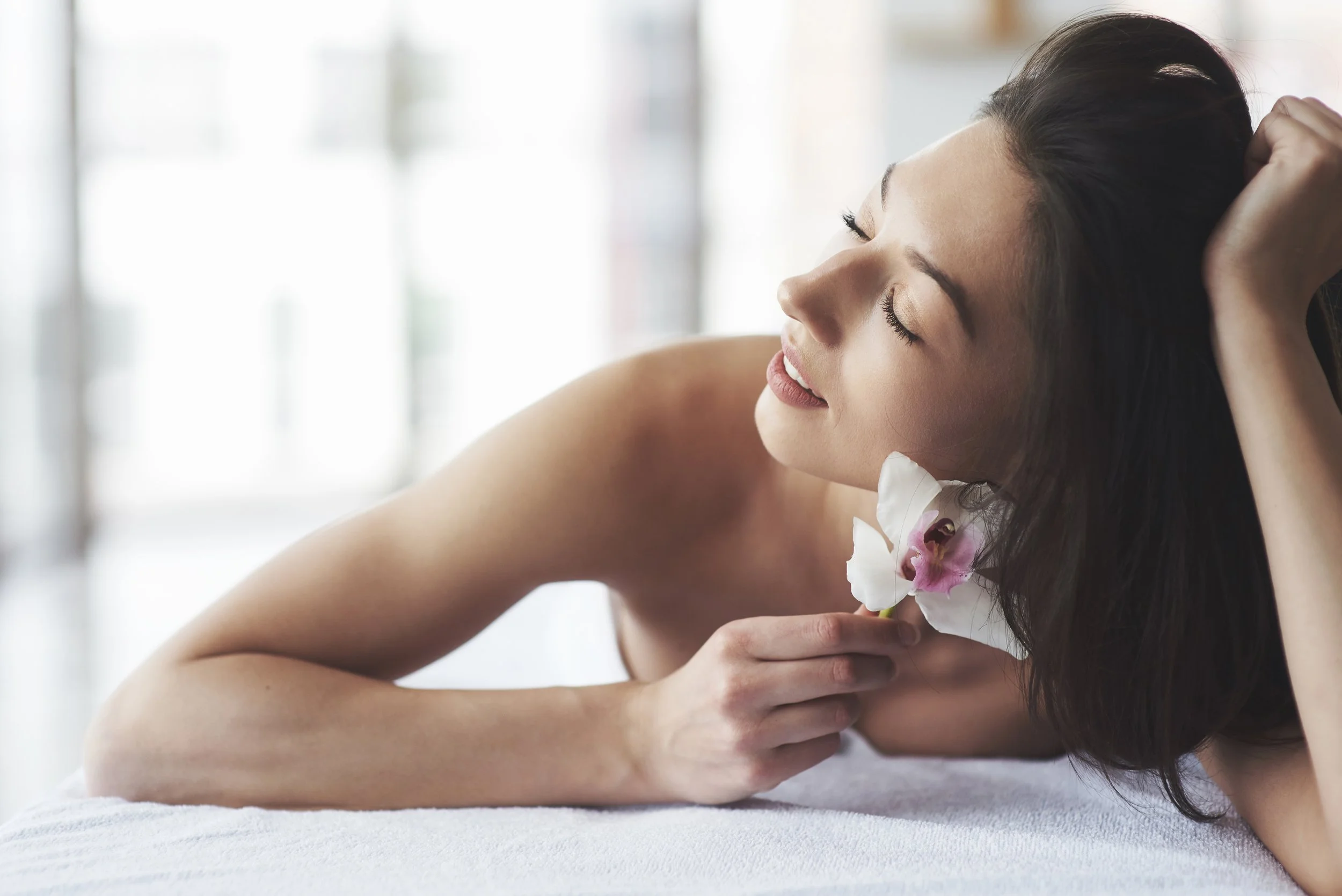 A woman with dark hair holding a white and pink flower close to her chest with her eyes closed and a relaxed expression, lying on a soft surface in a bright room.