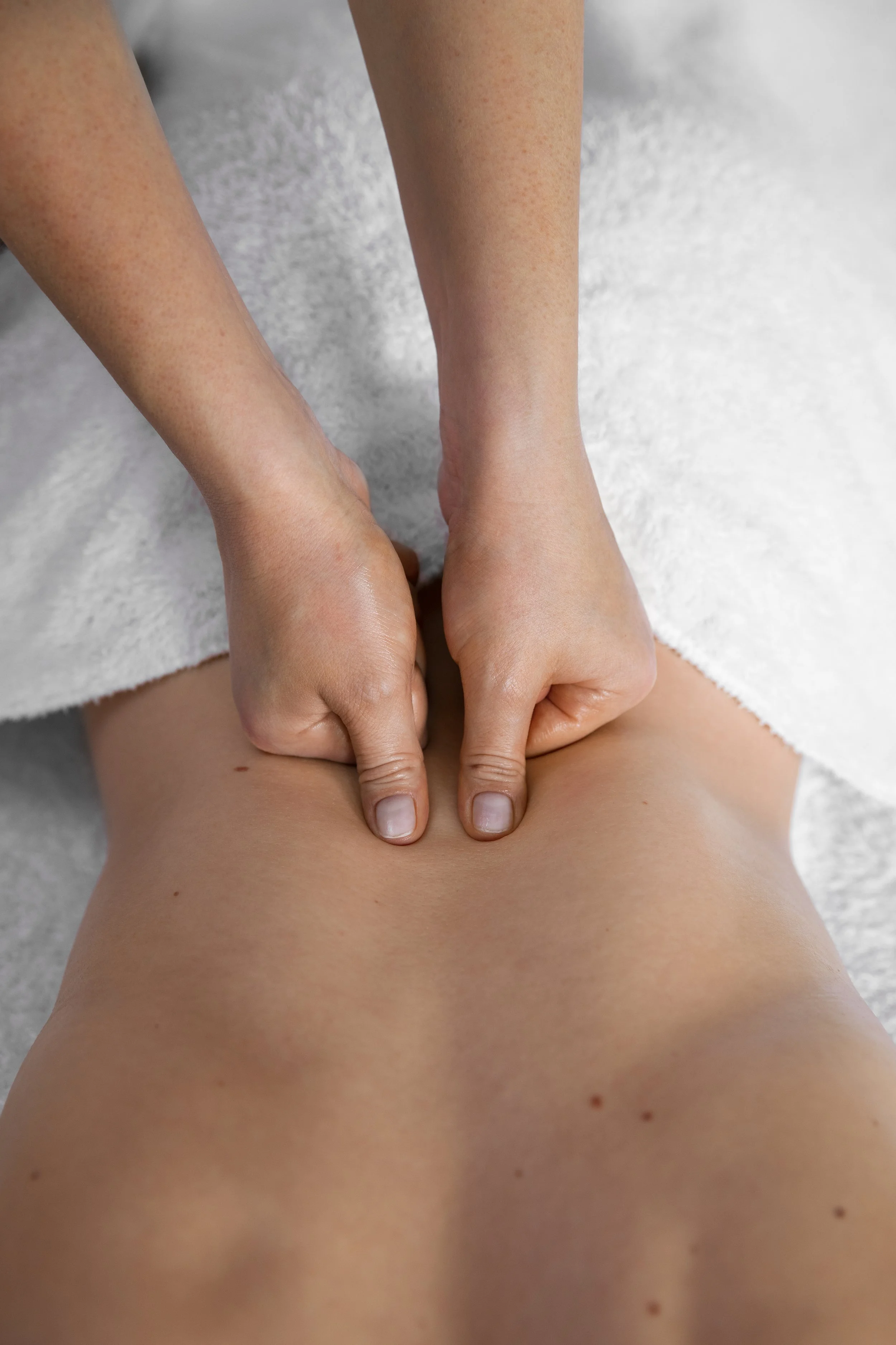 Person receiving a massage, lying face down on a massage table with a towel covering their lower body, while a therapist presses on their back.