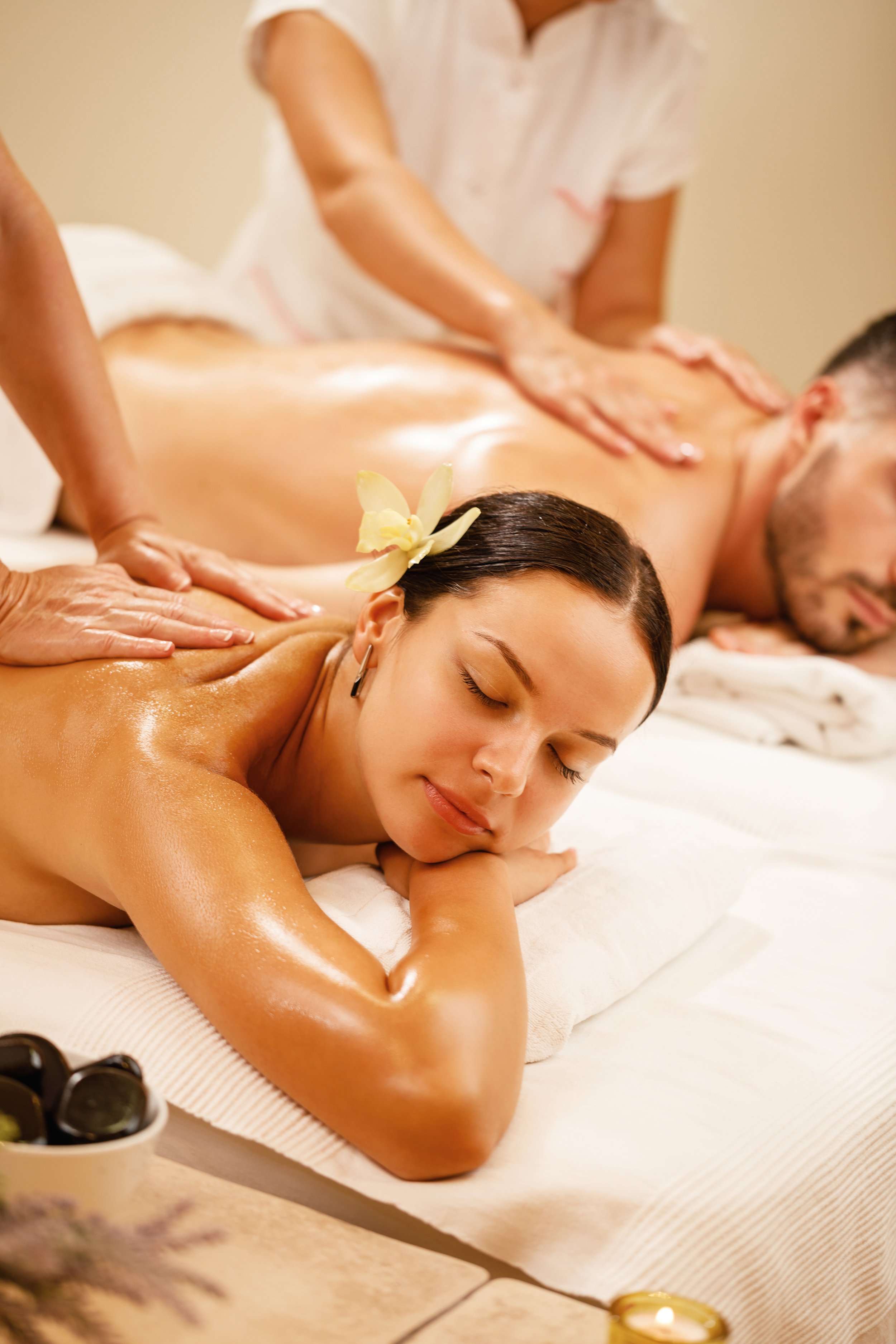 A woman with a flower in her hair is lying face down on a massage table, getting a massage. The massage therapist is applying pressure to her back. The scene appears to be in a spa or massage parlor.