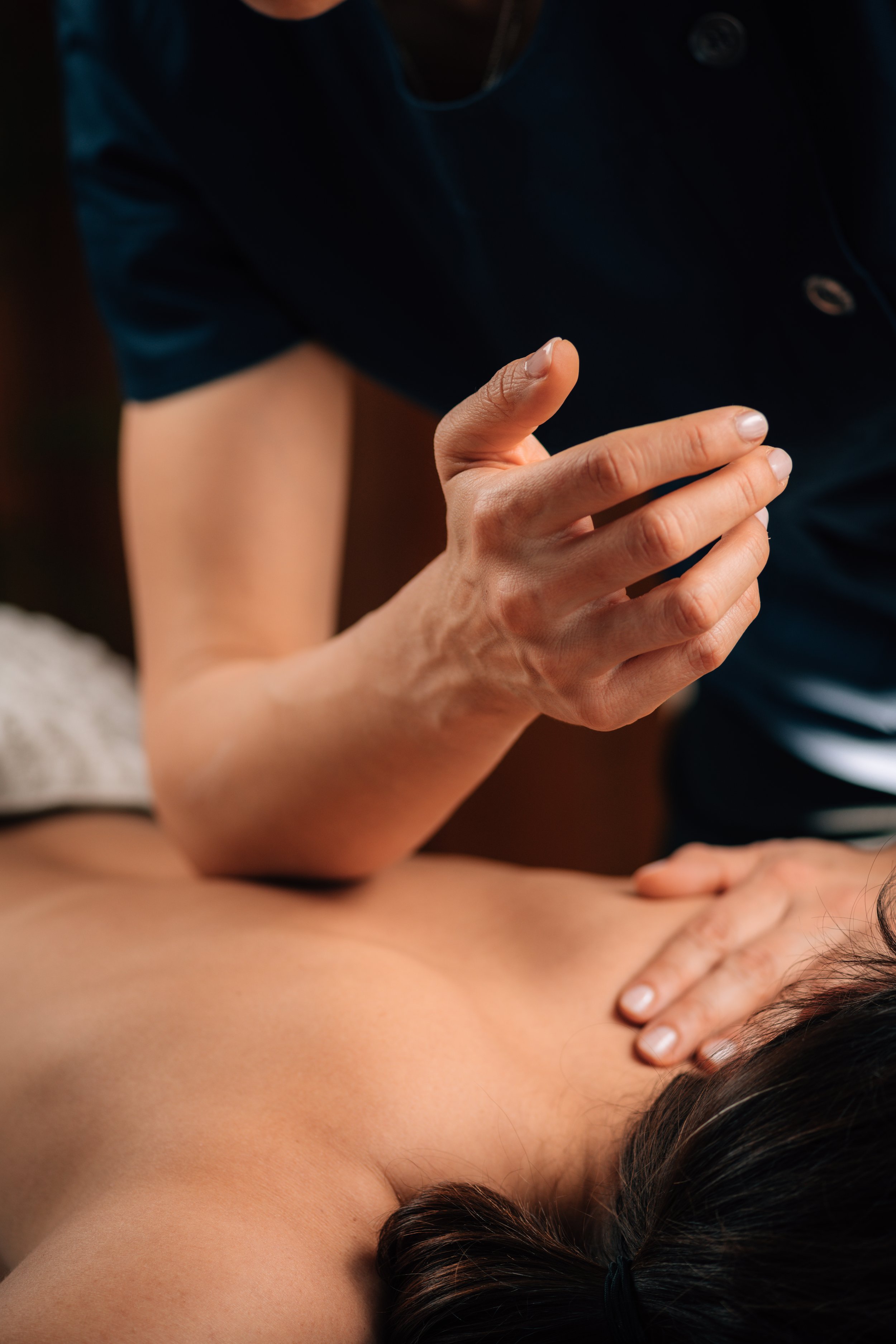 A woman lying on her stomach with her head turned to the side, receiving a massage on her back from a massage therapist. The therapist's hands are pressing on her back.