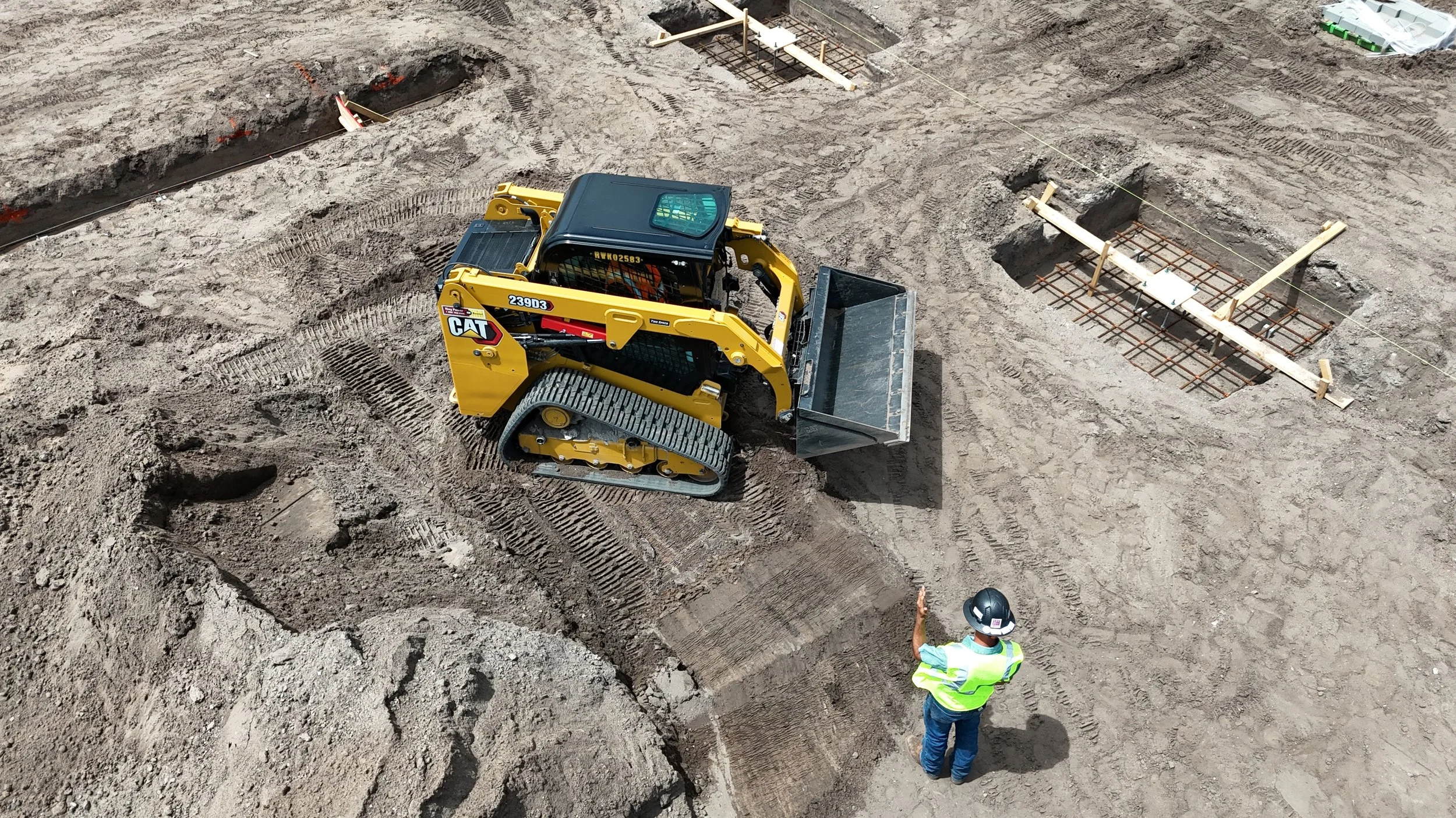 Construction site with a small yellow Caterpillar bulldozer and a construction worker wearing a safety helmet and vest, working near concrete foundation rebar.