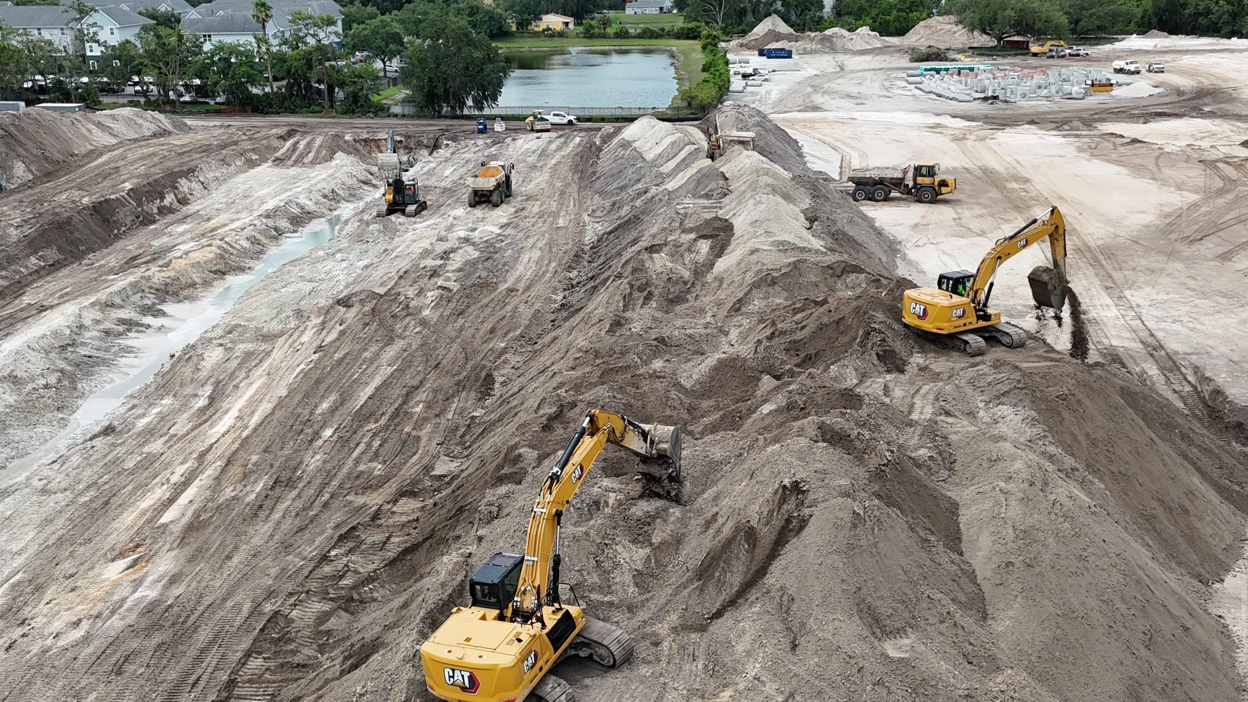 Construction site with multiple excavators leveling the dirt, with a small body of water and residential houses in the background.