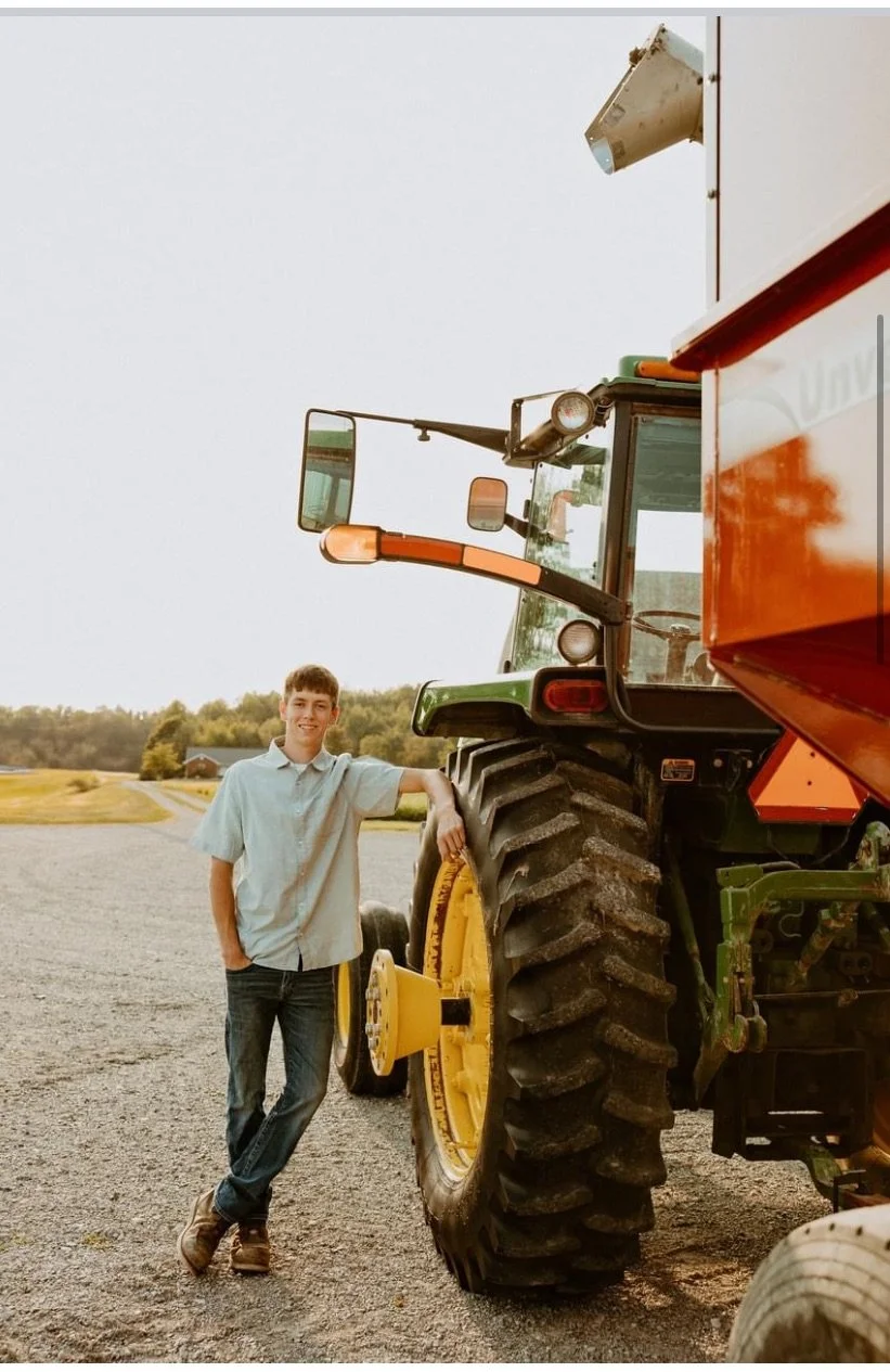 A young man standing next to a large green and yellow tractor outdoors on a gravel surface, with fields and trees in the background.