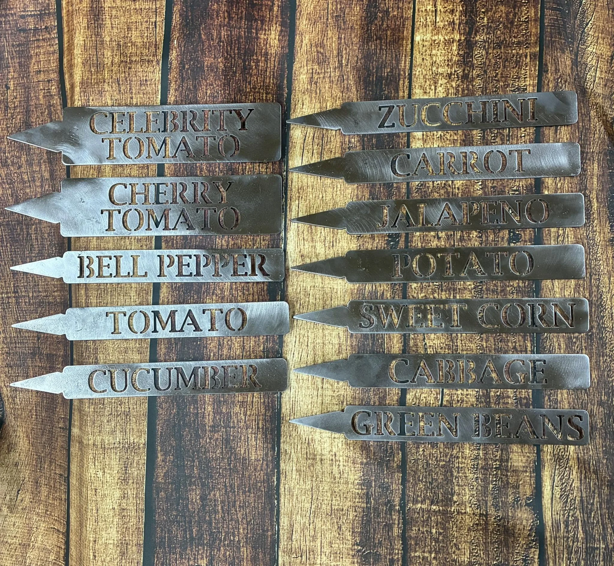 Metal plant markers with cut-out words, labeled for different vegetables and herbs, arranged on a rustic wooden surface.