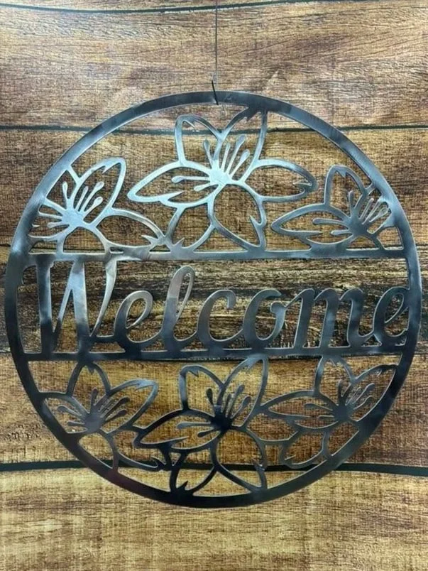 Decorative metal wall hanging with a circular design featuring flowers and the word "Welcome" in the center, mounted on a wooden background.