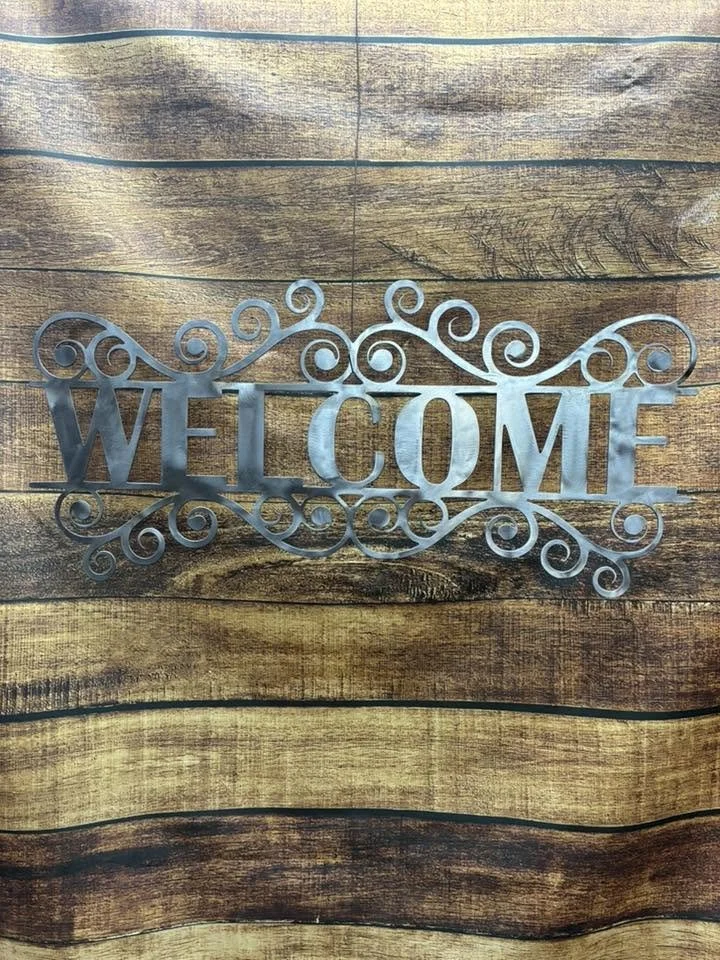 Metal sign with the word 'WELCOME' surrounded by decorative swirls, placed on a wooden surface.