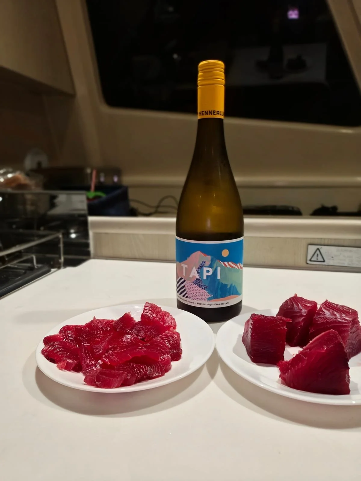 A bottle of TAPI cider between two plates of raw tuna sashimi on a white counter, with kitchen appliances and a window in the background.