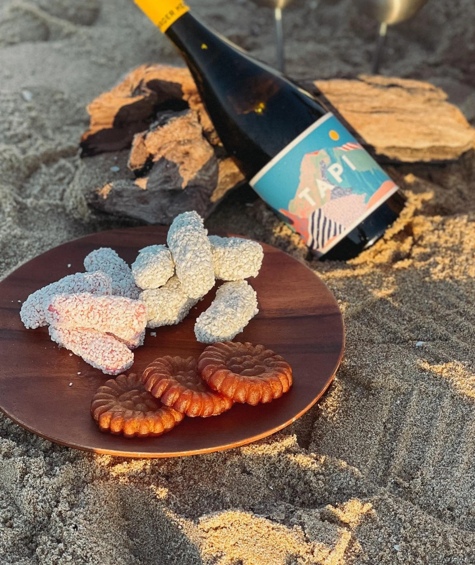 A wooden plate with white cinnamon-sugar-coated pretzels, some chocolate cookies, and another type of cookie on a sandy surface. In the background, there's a bottle of Tampico juice and a few small umbrellas.