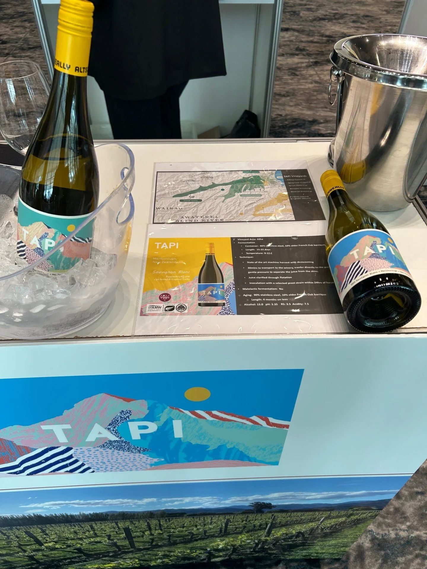 A display for TAPI vineyards featuring two bottles of white wine, a glass, a stainless steel ice bucket, and informational signage about the vineyard and wine production.