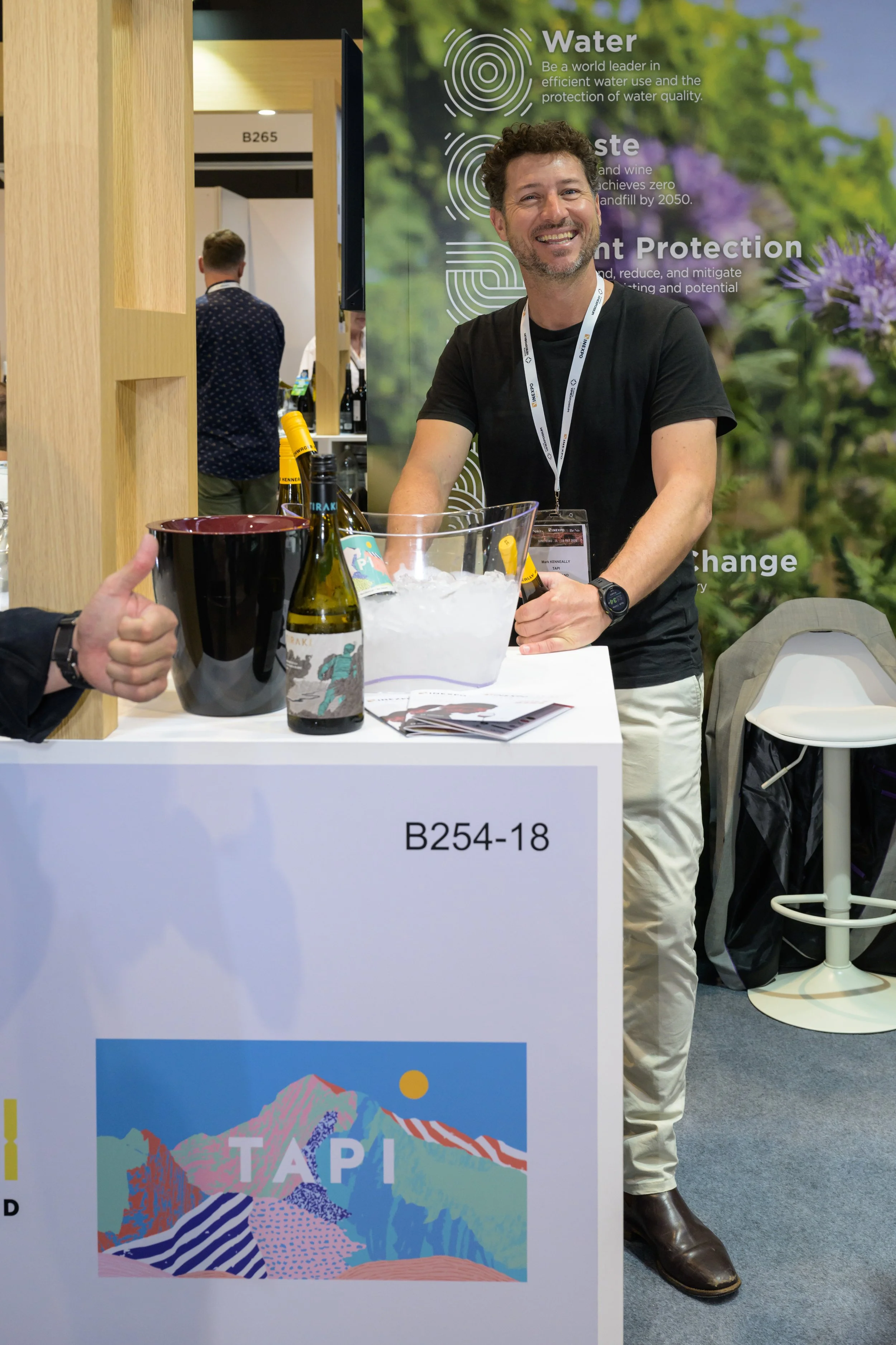 A man with curly hair and a beard, smiling, standing at a booth with a tropical landscape poster featuring mountains, trees, and the word "TAPI". He wears a black T-shirt, white pants, a wristwatch, and a lanyard with an ID badge. There are bottles o