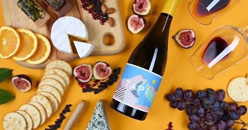 A bottle of wine surrounded by cheese, crackers, grapes, figs, oranges, and snacks on a yellow table.