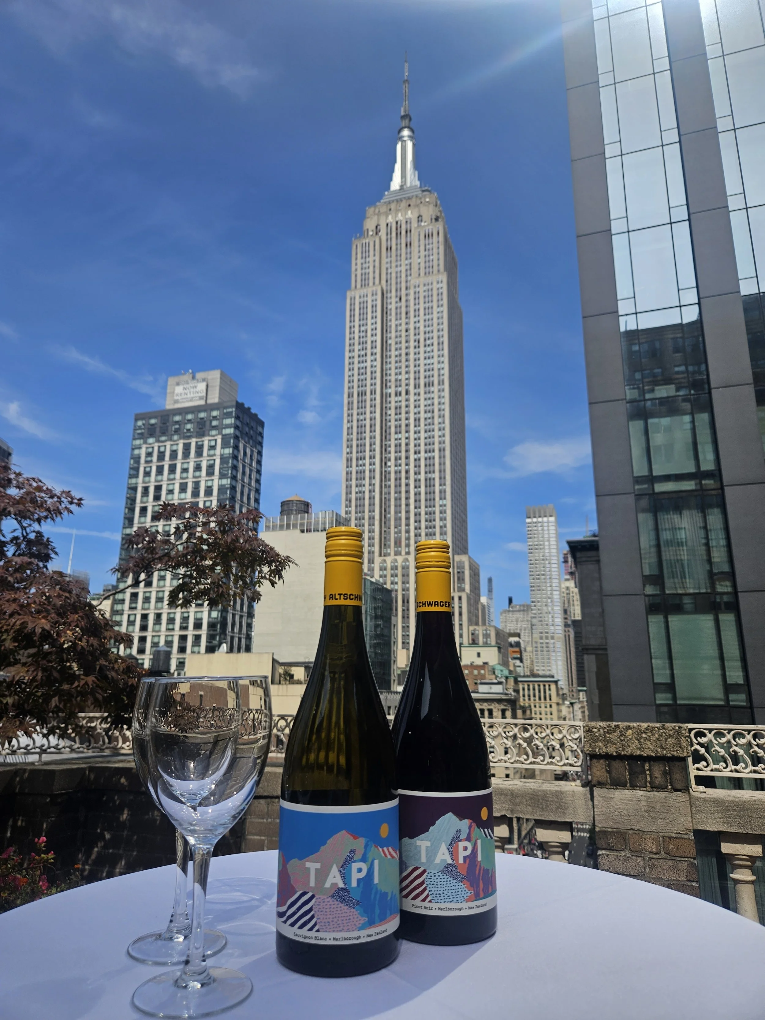 On a white table, there are two wine bottles with colorful labels that read 'TAPI', a clear wine glass, and a view of the Empire State Building against a blue sky with some clouds.