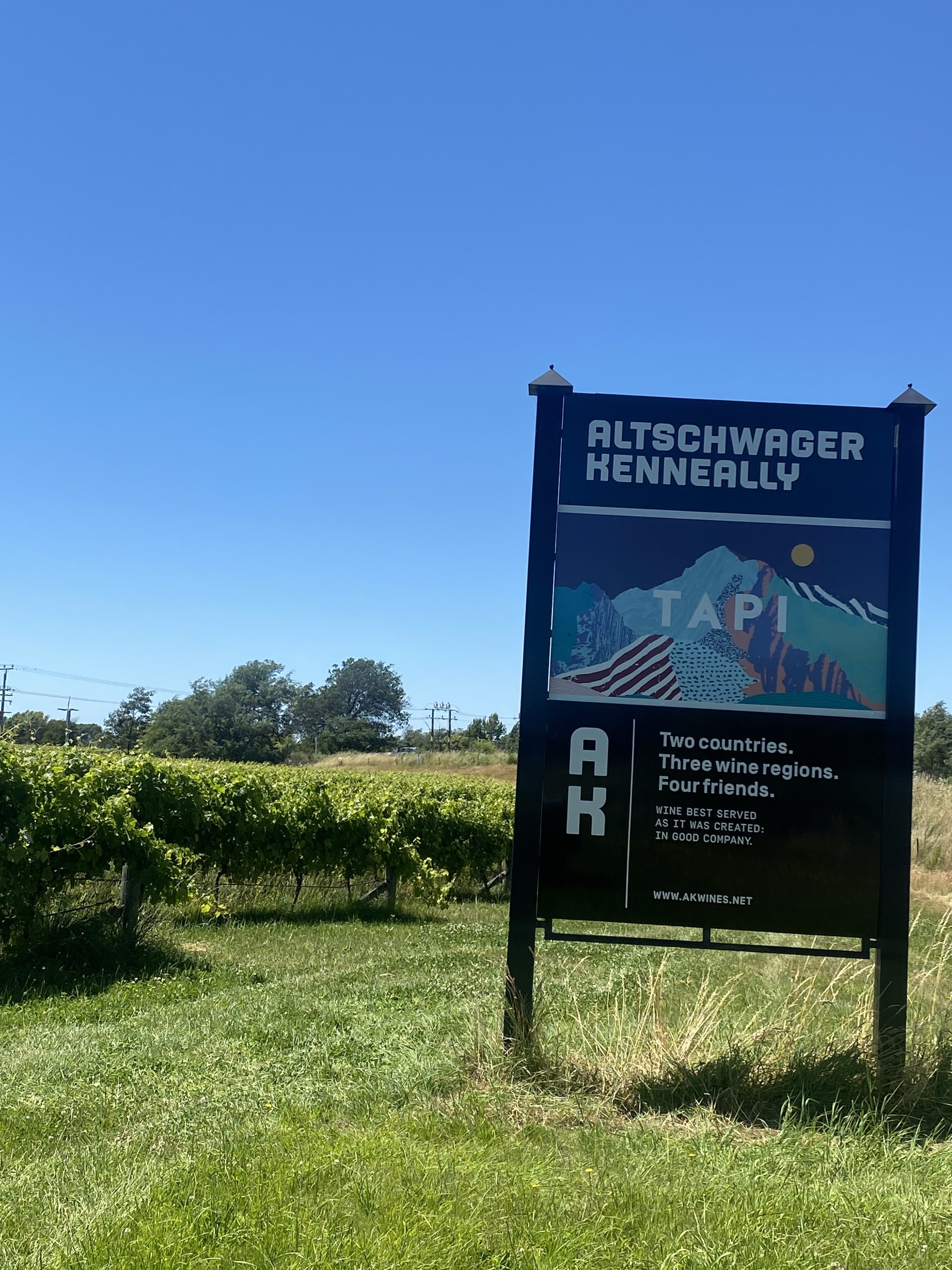 Sign for AltSchwager Renneally winery with a mountain landscape illustration, situated in a green field with trees and power lines in the background under a clear blue sky.