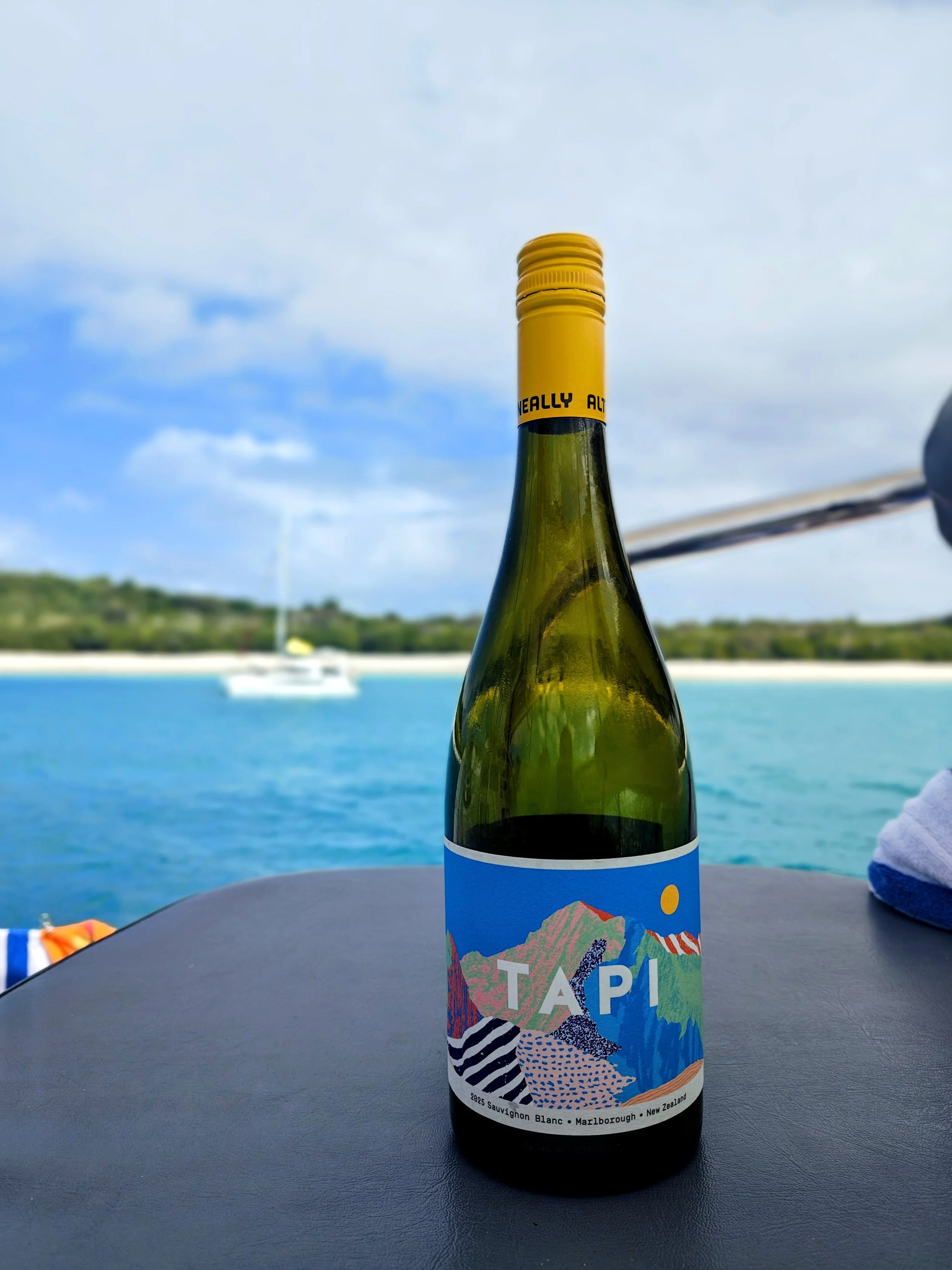 A bottle of Sauvignon Blanc wine with a colorful label featuring mountains and abstract patterns, placed on a table outdoors near a body of water with a boat and hills in the background under a partly cloudy sky.