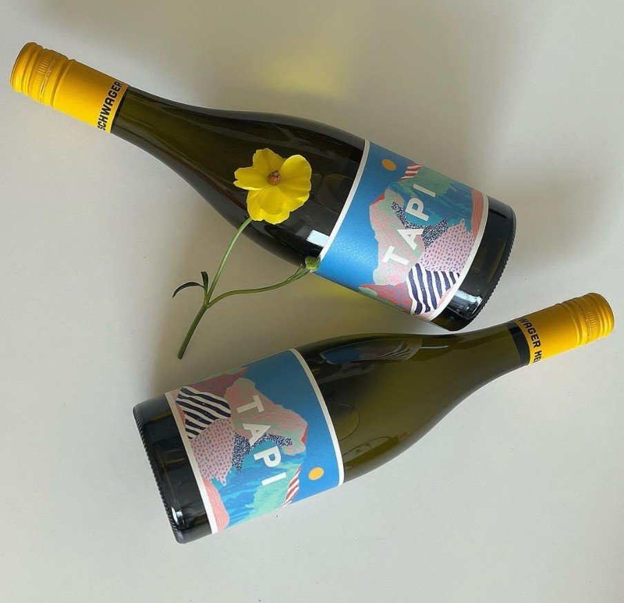 Two wine bottles with colorful landscape labels, arranged on a white surface, with a yellow flower placed beside one bottle.