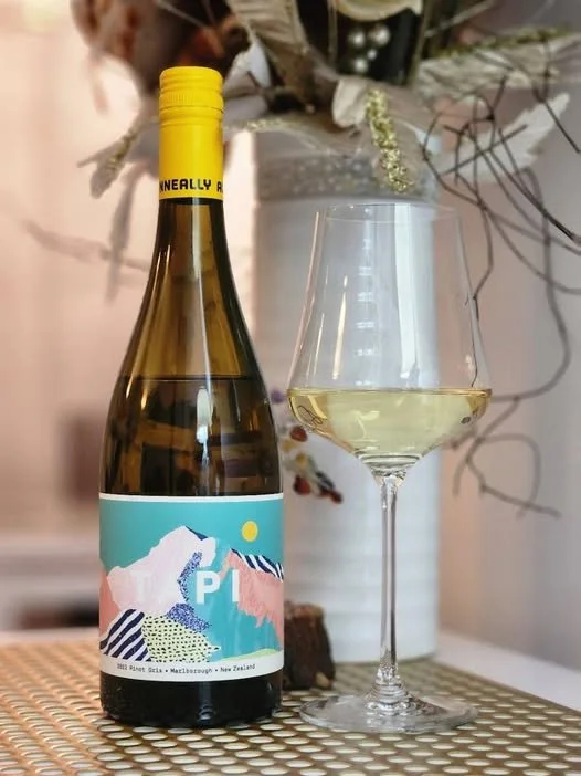 A bottle of white wine with a blue and pink mountain label next to a filled wine glass, placed on a woven surface with a decorative vase and dried flowers in the background.