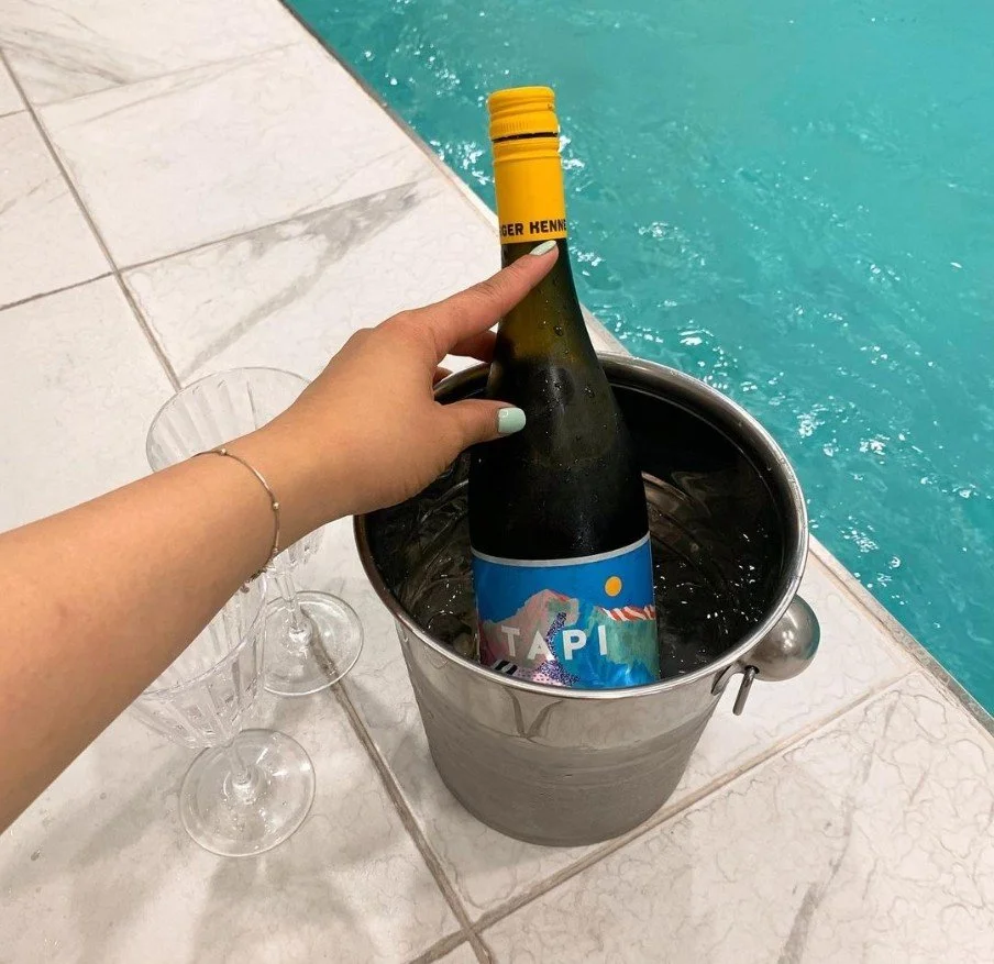 A person is placing a bottled beverage into a metal ice bucket by a swimming pool.