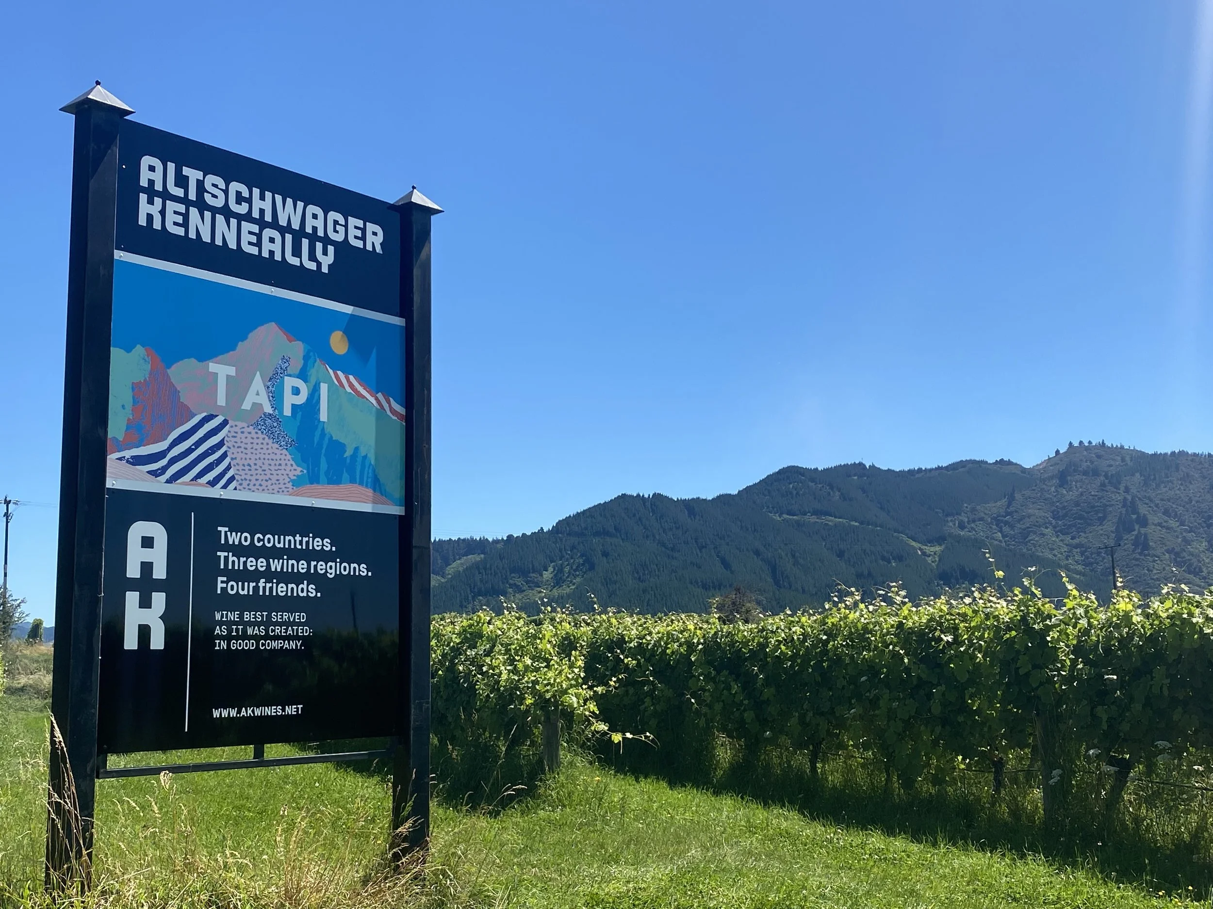 Sign welcoming visitors to Altenweger Kenneally, featuring a colorful mountain landscape illustration, against a background of green hills and blue sky.