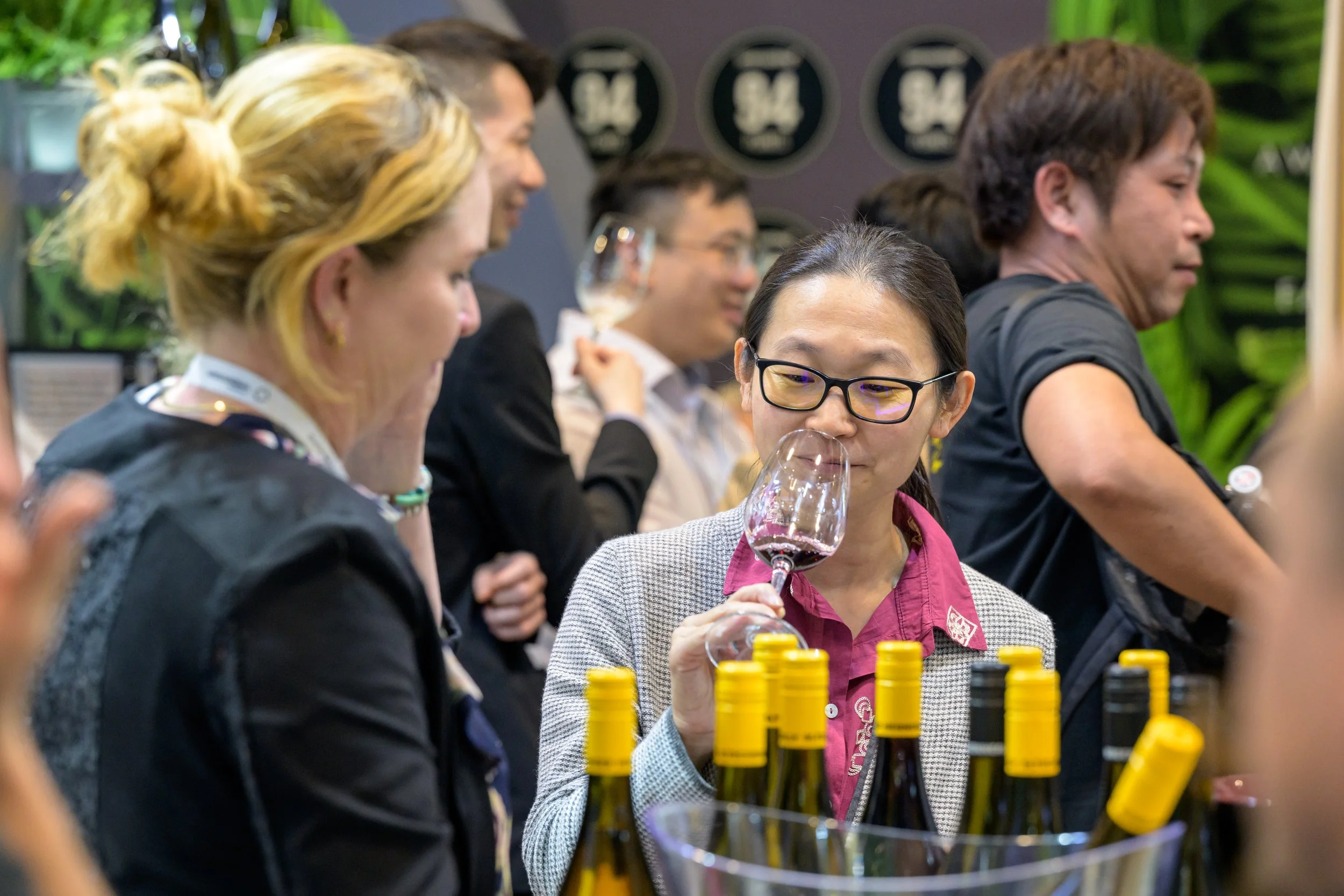 A group of people at a wine tasting event, some tasting and smelling wine, with wine bottles and bottles caps on the table in the foreground.