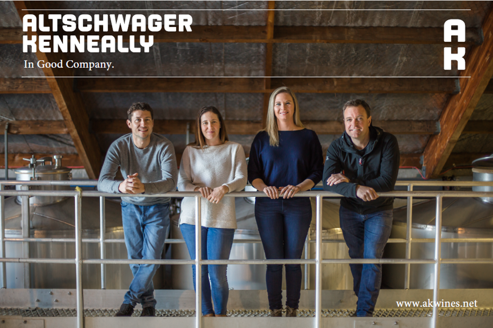Four people leaning on a metal railing inside a winery or brewery building with wooden beams and metal tanks in the background.