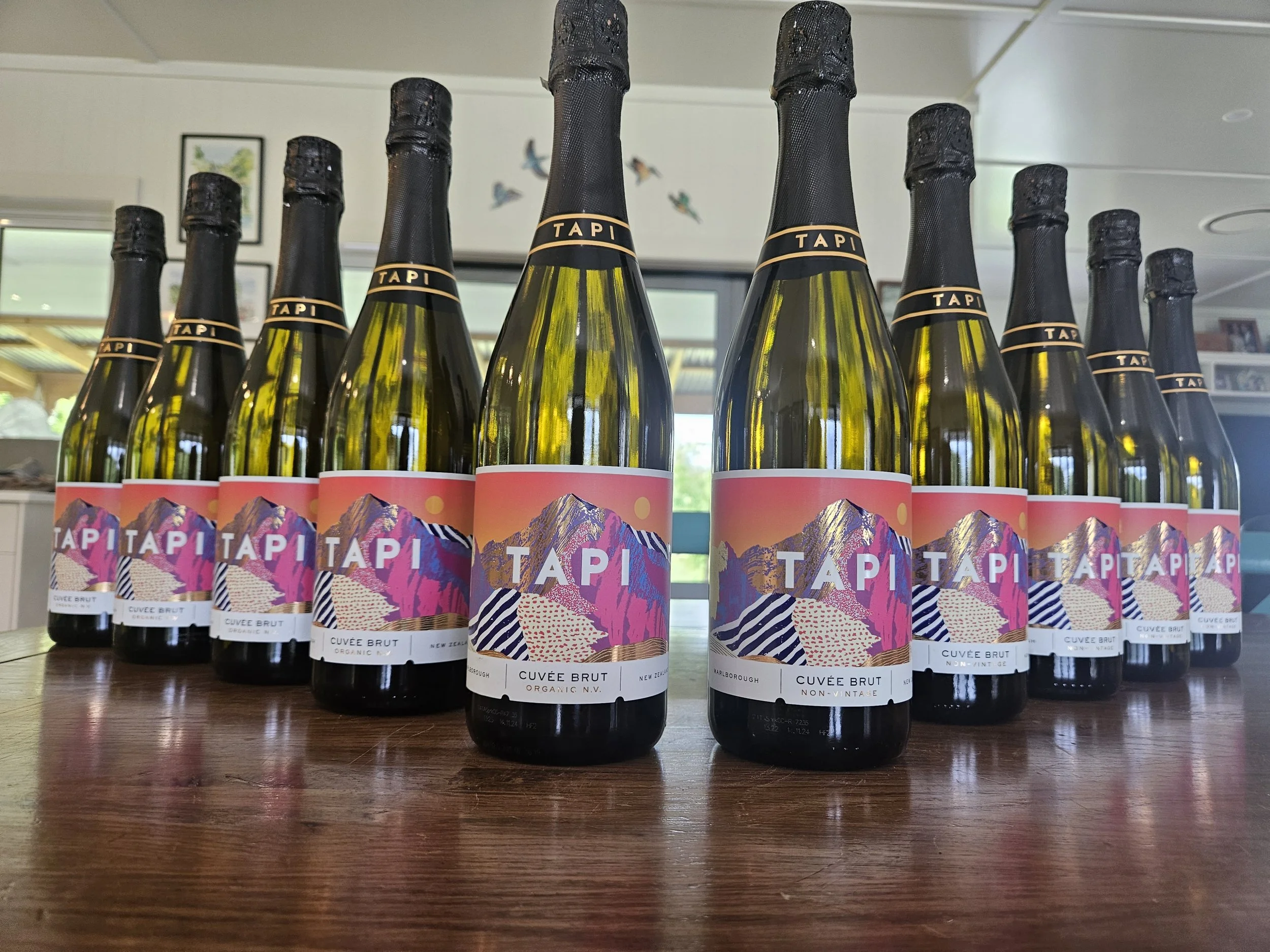 A row of bottles of sparkling wine with colorful label art featuring a mountain scene, arranged on a wooden surface indoors, with windows showing greenery in the background.