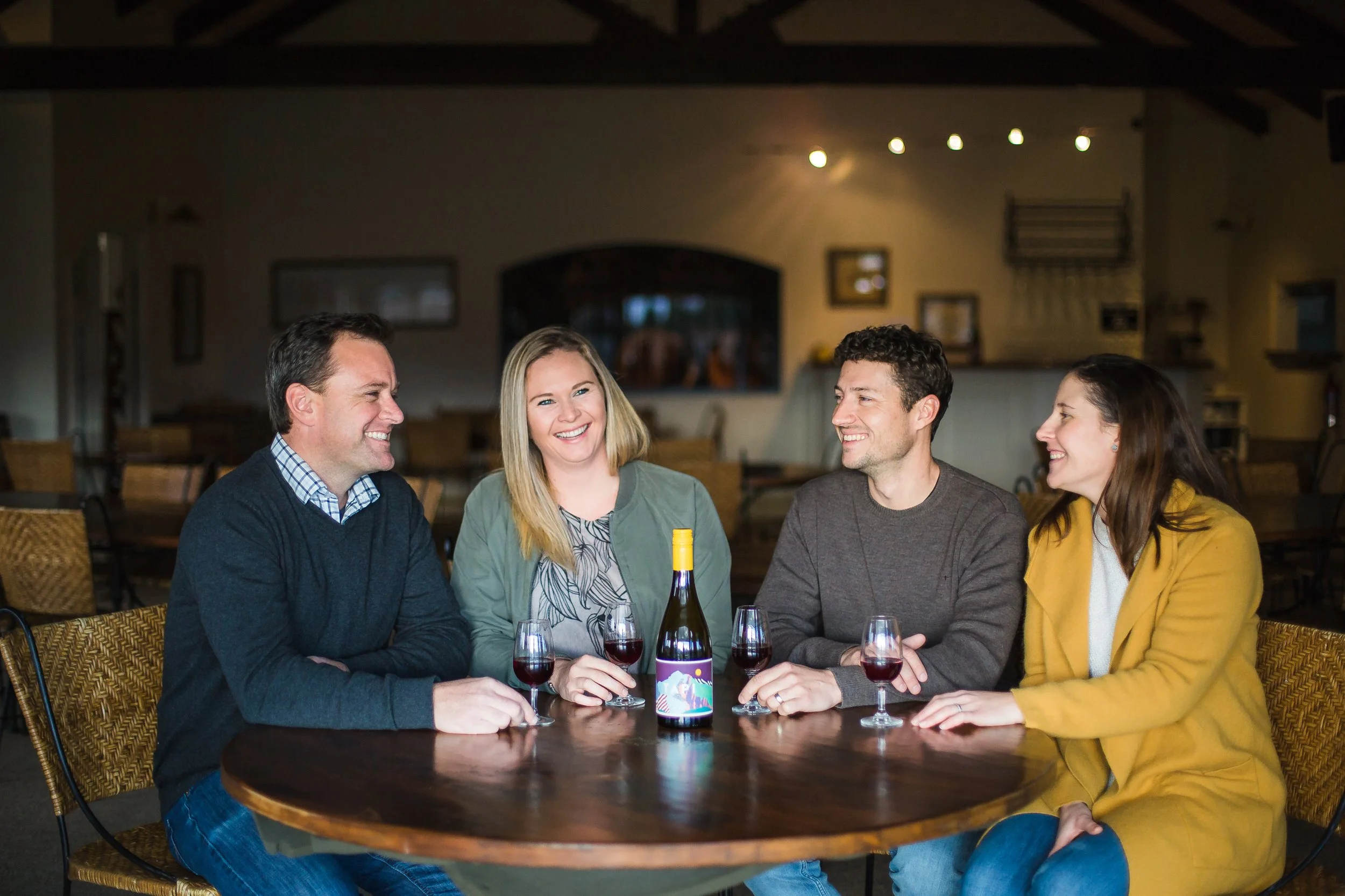 Four friends sitting around a table in a restaurant, smiling and talking. There are four glasses of red wine, a bottle of wine, and an empty wine glass on the table.