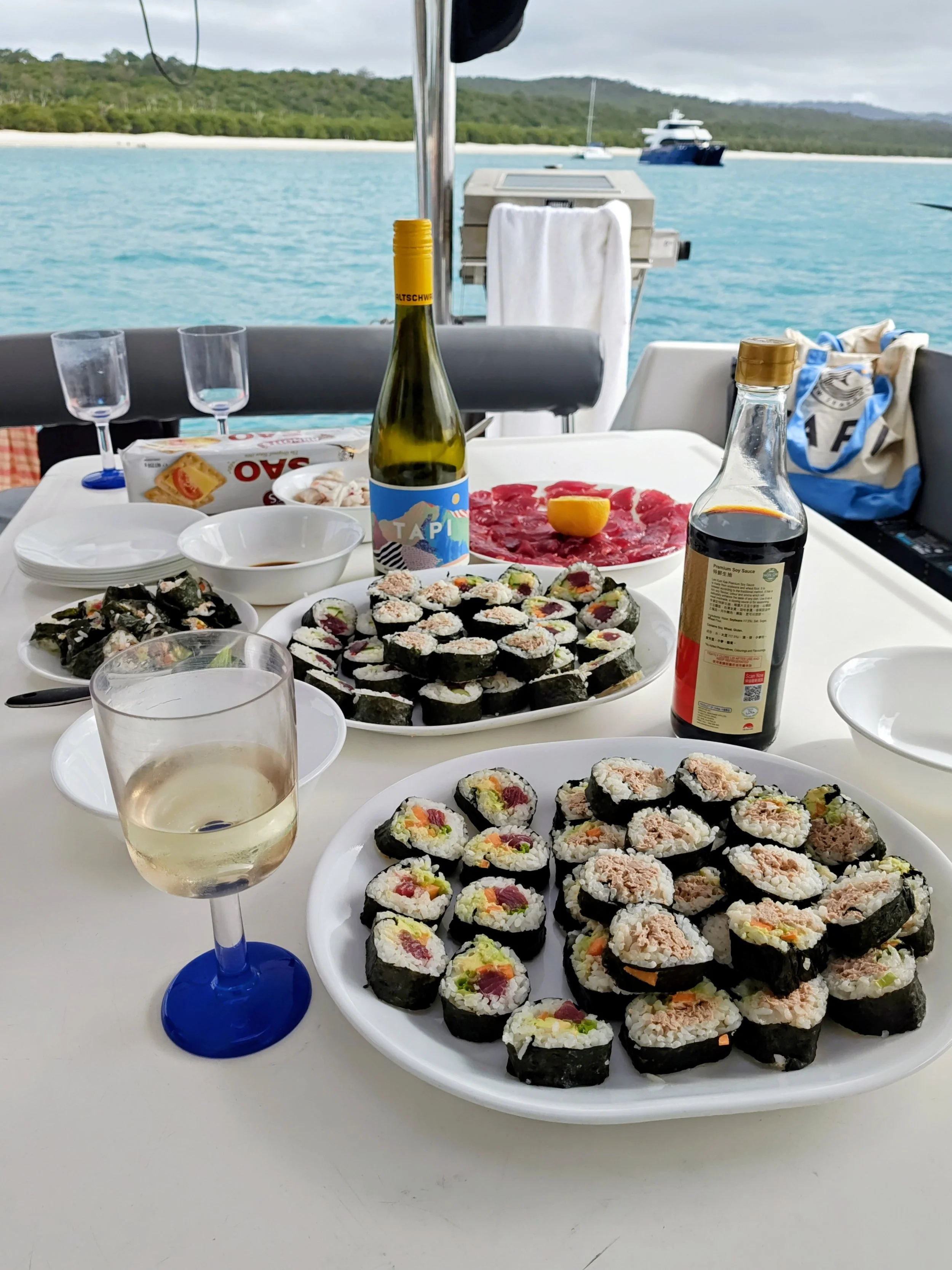 A table on a boat with various sushi rolls, a glass of white wine, soy sauce, and other condiments. In the background, water, a yacht, and a shoreline with trees are visible.