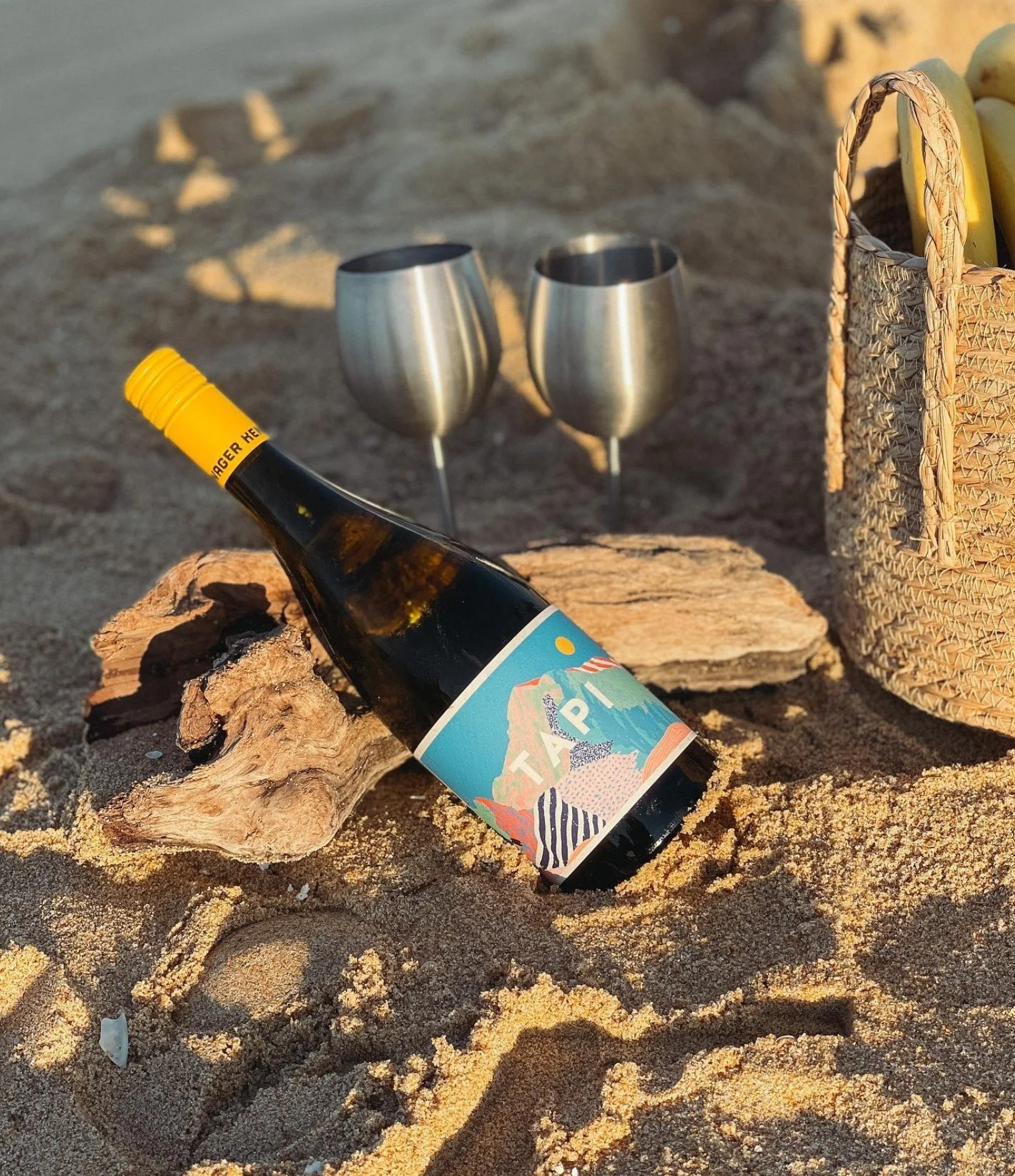 A bottle of sparkling wine with a colorful label resting on a piece of driftwood on a sandy beach. In the background, two metal wine glasses and a woven basket with yellow and green objects are visible.