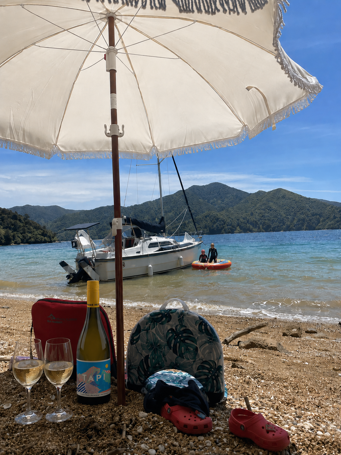 A beach scene with a large white umbrella, two glasses of white wine, a bottle of wine, a backpack, a small patterned tent, red Crocs, and an inflatable boat on the sand. In the background, there is a boat and two people in the water, with green moun