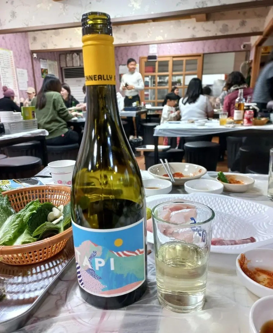 A bottle of white wine with a glass of wine on a table set for a meal. The table has various dishes, including lettuce and small bowls of food. In the background, people are sitting at tables in a lively restaurant.