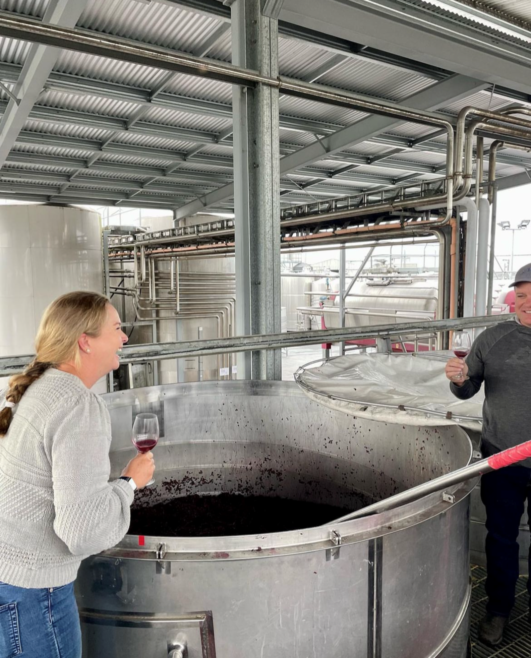 Two people tasting red wine from glasses near large stainless steel fermentation tanks in a winery.
