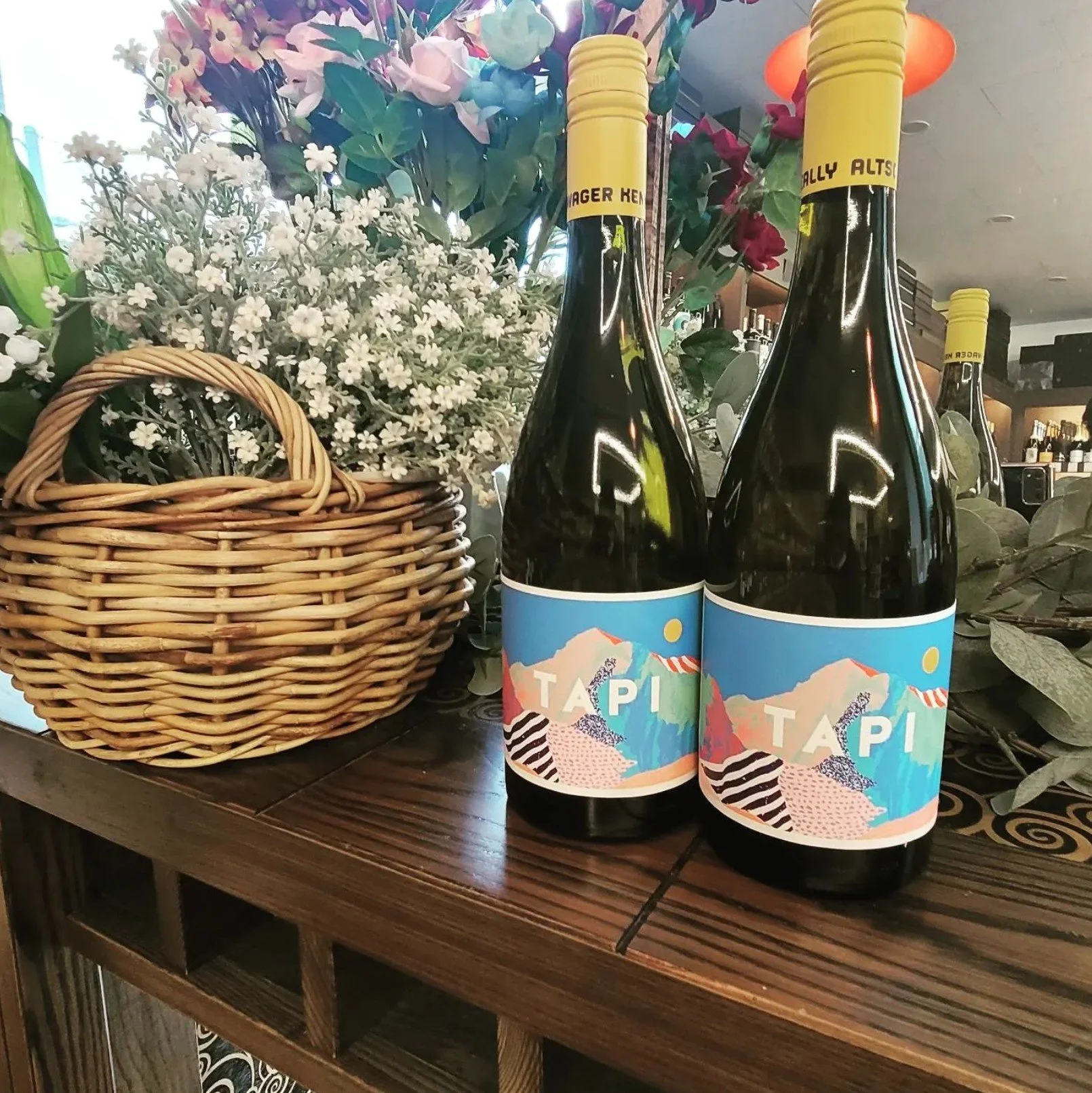 Two bottles of tap water with colorful mountain and landscape labels are placed on a wooden table next to a wicker basket and a floral arrangement with white, pink, and purple flowers. The background shows a cozy indoor setting with warm lighting.