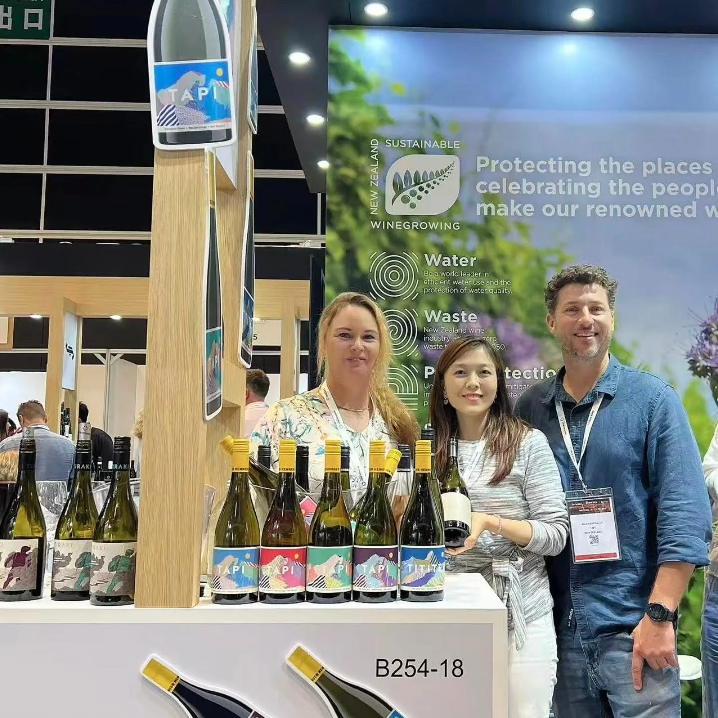 Three people, two women and one man, standing behind a table displaying bottles of wine at an indoor event. There is a large promotional poster in the background with text about New Zealand wine, sustainability, water, waste, and protection.