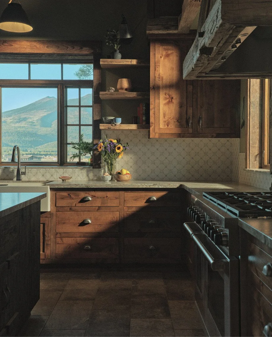 A rustic kitchen with wooden cabinets, a window showing a mountain landscape, a vase of wildflowers, and a stove.