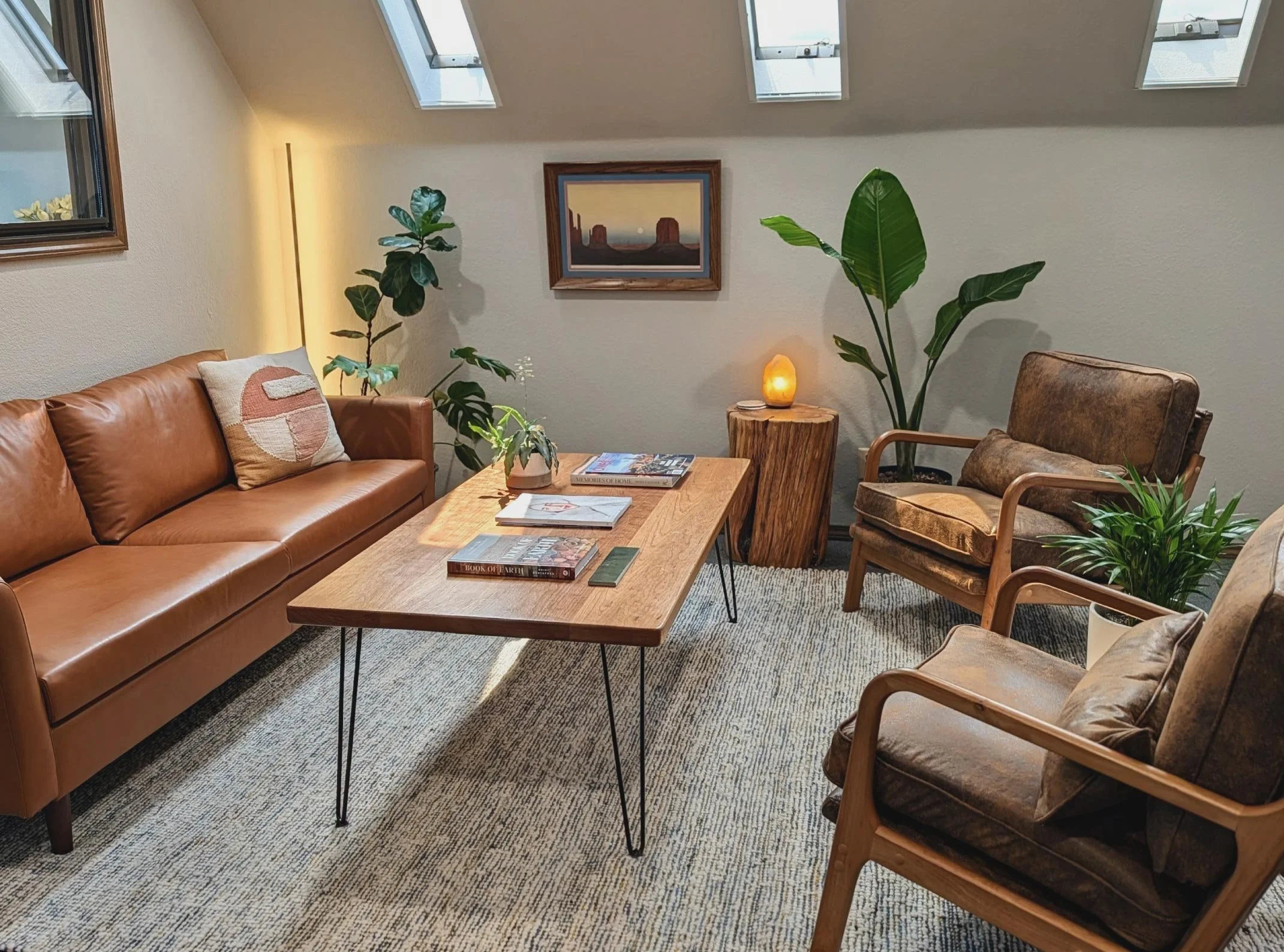 A cozy living room with a tan leather sofa, a wooden coffee table with magazines, and a chair with a wooden frame and brown upholstery, decorated with plants.