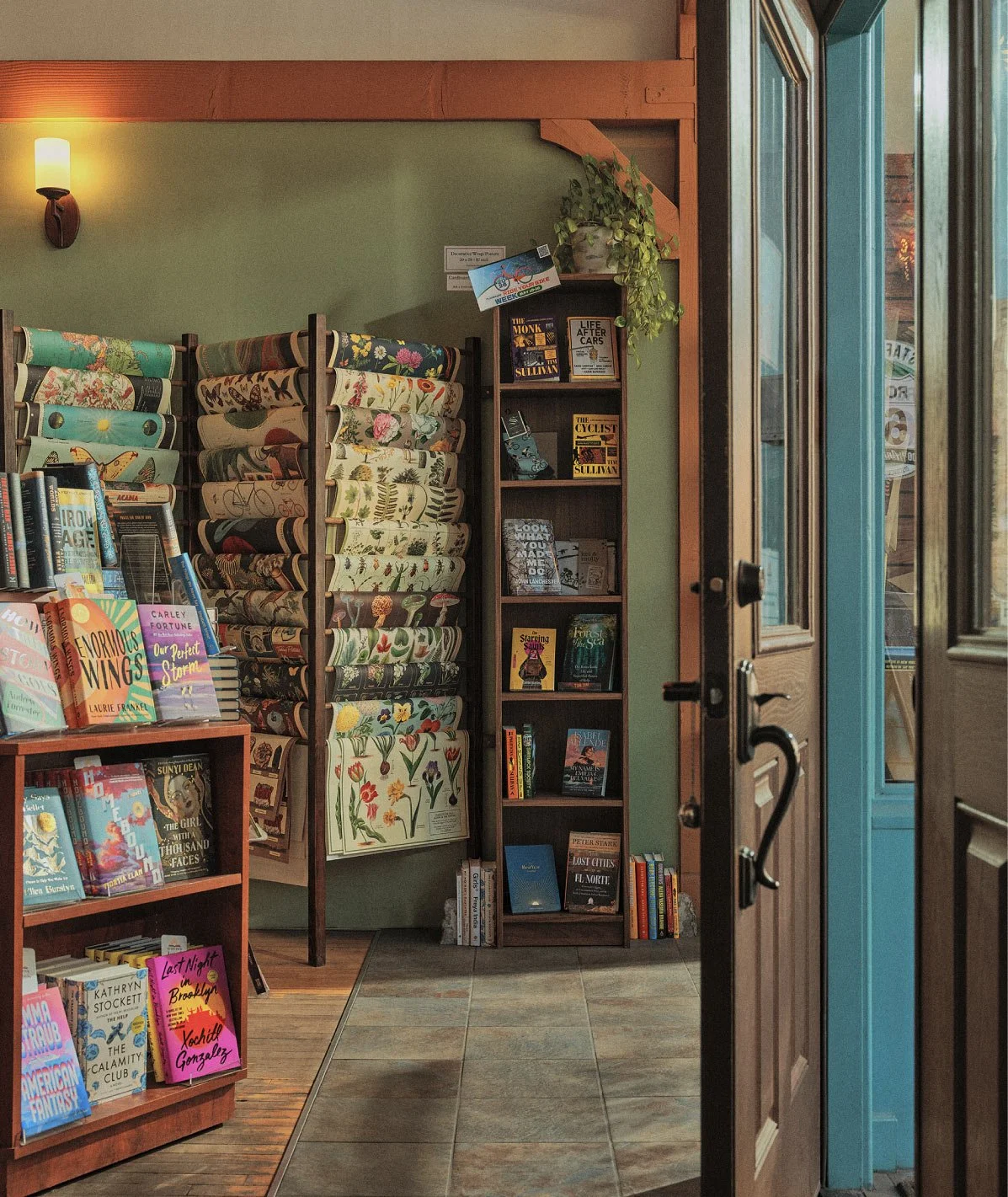 Inside a cozy bookstore with shelves of books, a display rack of colorful fabric prints, and a bookshelf filled with various books. The store has a warm ambiance with a wall-mounted light and large glass door letting in natural light.