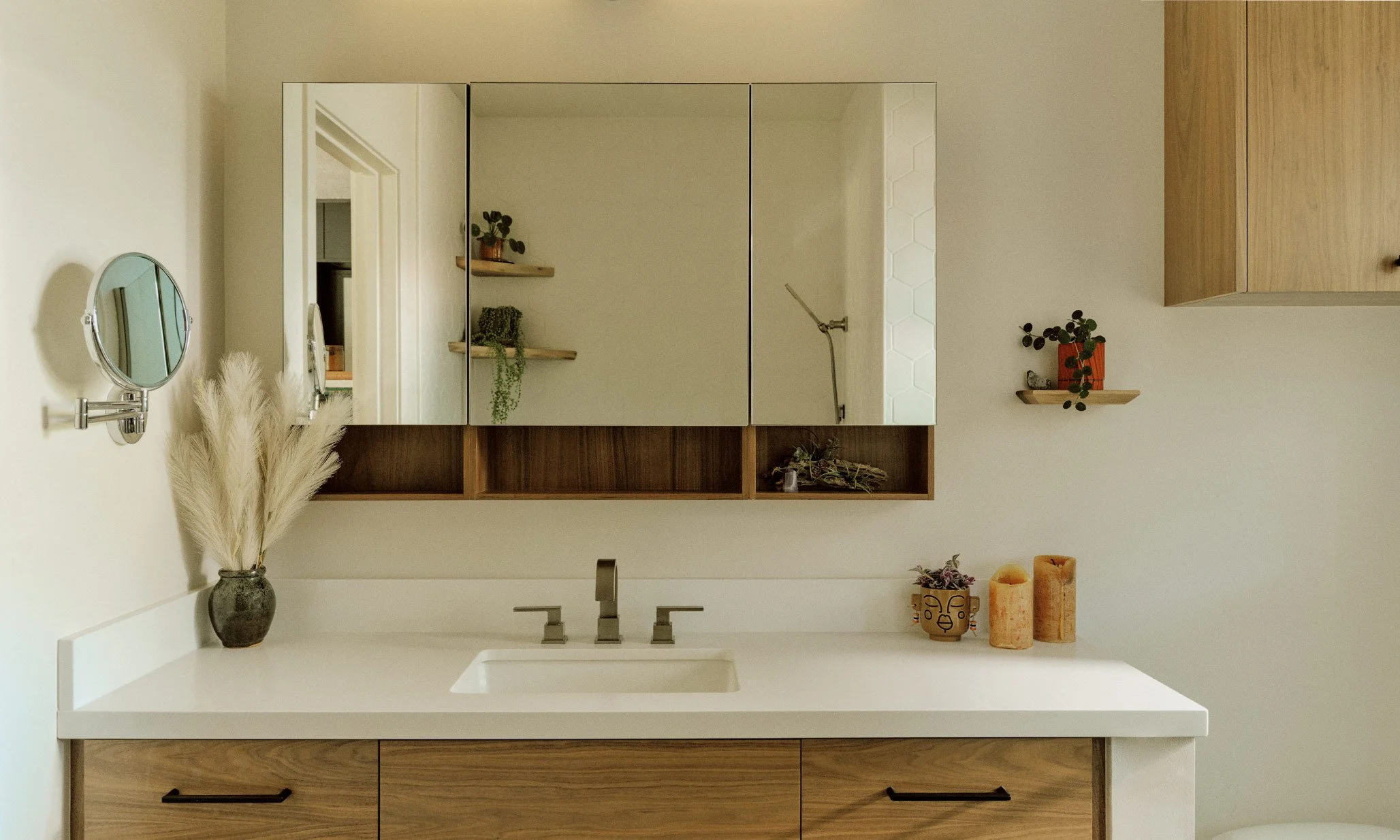 A modern bathroom vanity with a white countertop, a built-in sink, and wooden cabinetry. Above the sink are three mirrored cabinets, and there are small decorative plants and candles on the countertop and shelves.