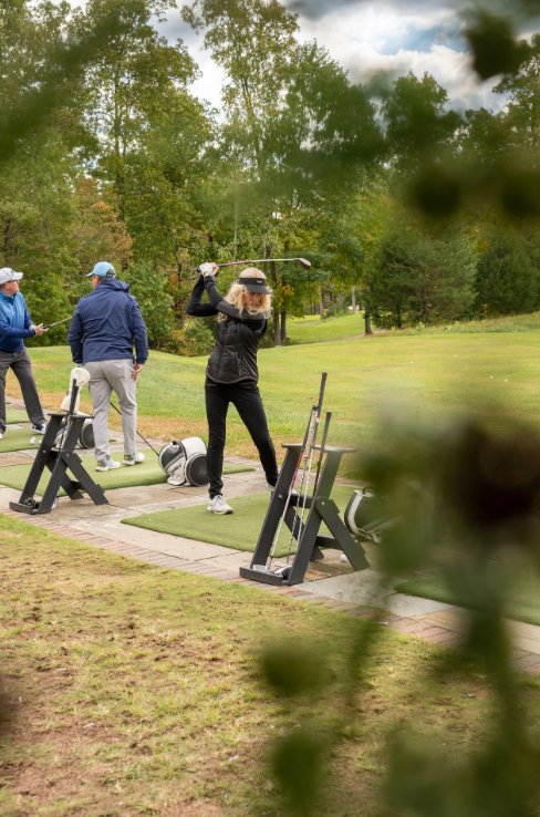 A woman swinging a golf club at a driving range with other golfers in the background.