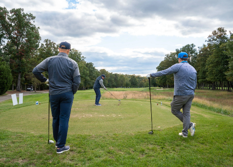 Three men playing golf on a course, with one preparing to swing and two others watching on a cloudy day.