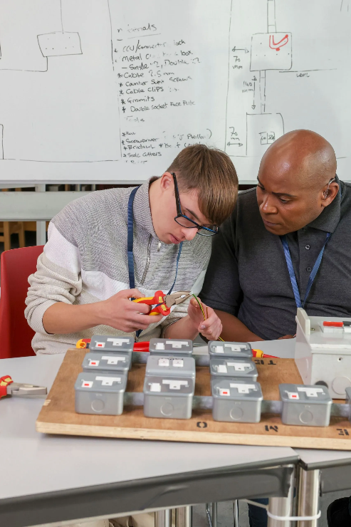 A young man and an older man work together on an electrical project with multiple circuit boxes on a table, in a classroom or workshop setting with whiteboard notes in the background.