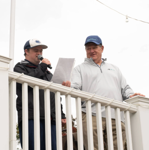Two men standing on a balcony, one holding a microphone and reading from a paper, the other listening, outdoors with an overcast sky.
